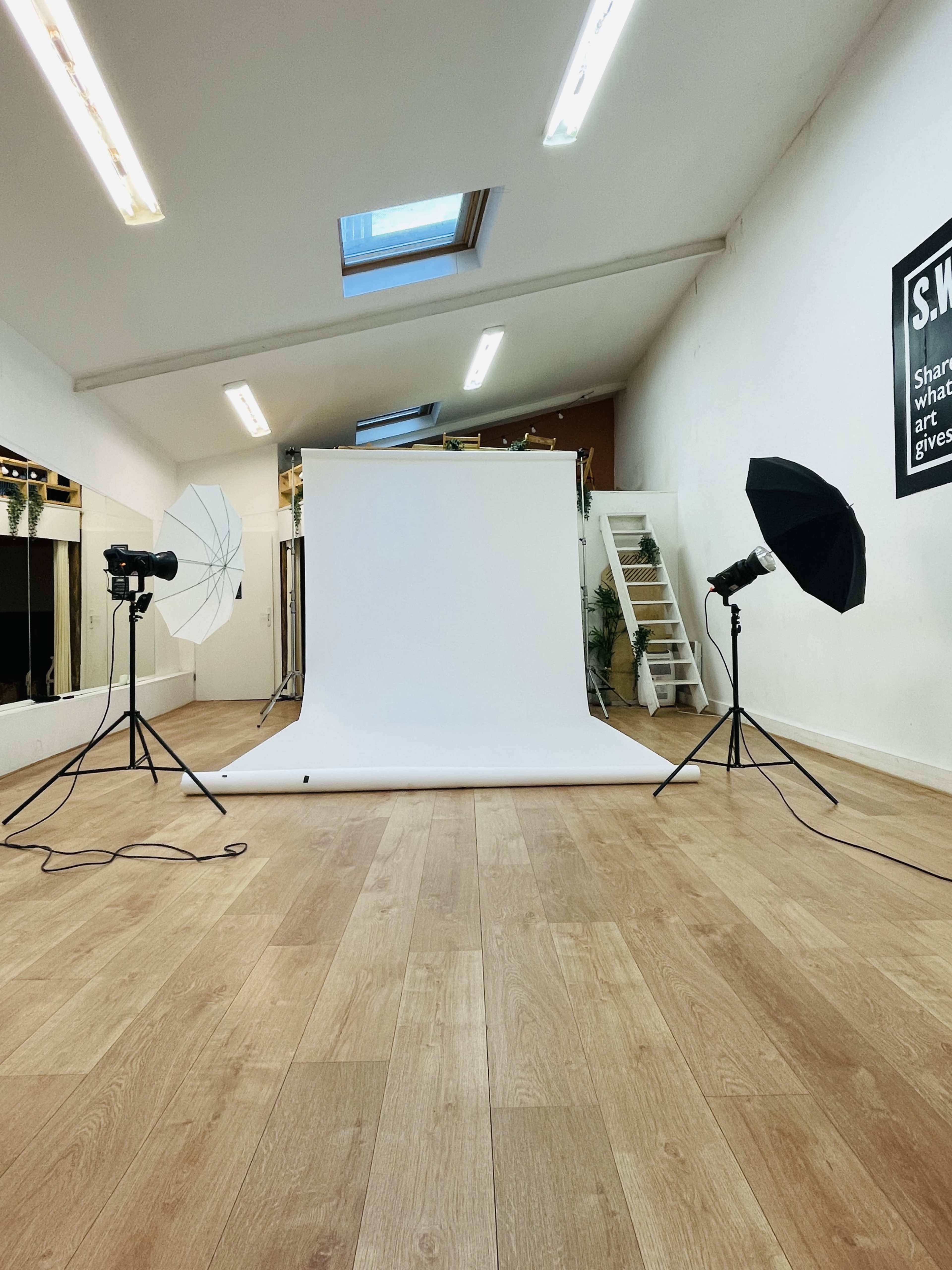 A photography studio features a white backdrop with lighting equipment set up on either side, along with a staircase and mirrors in the background.