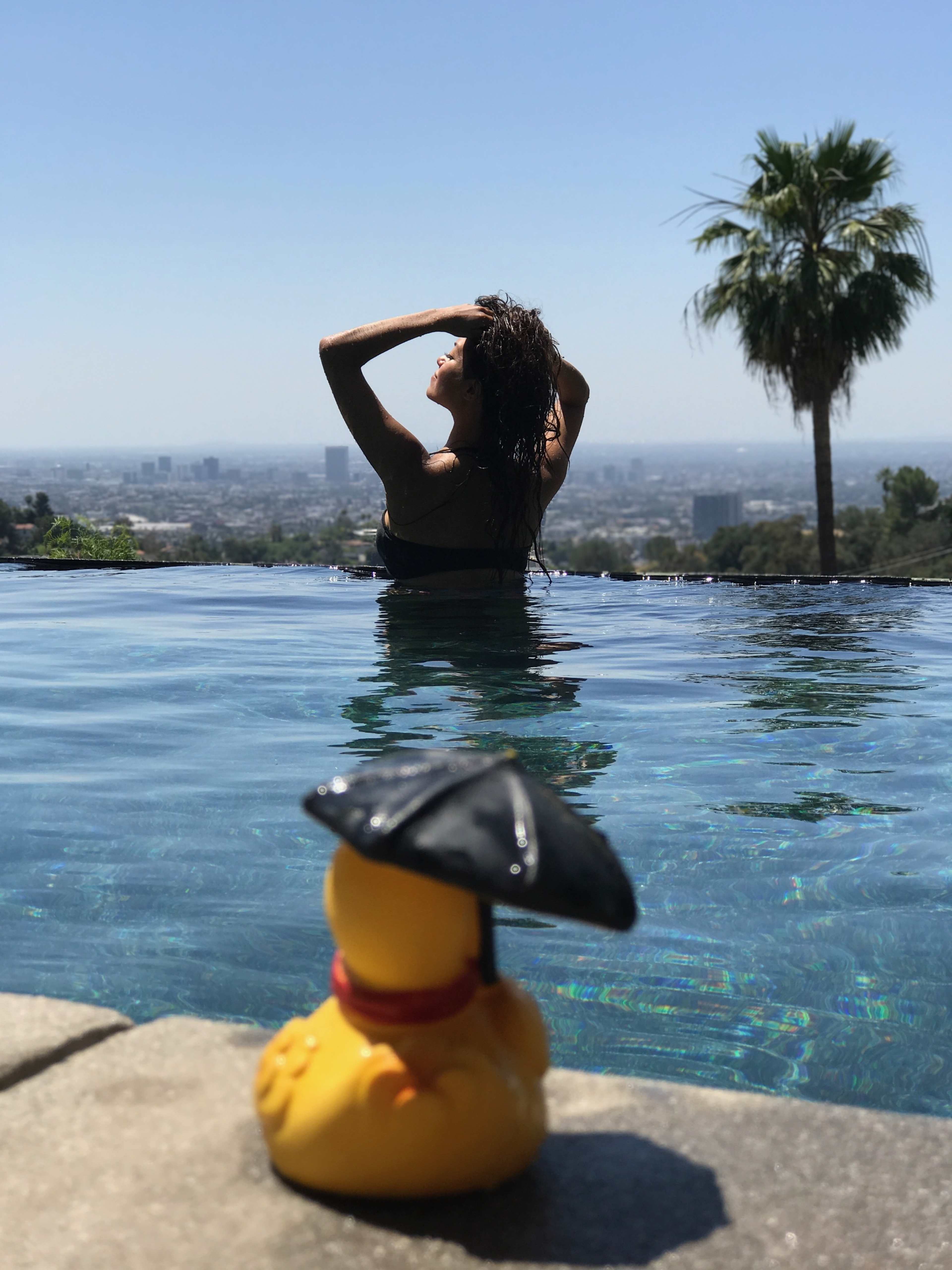 A woman stands in an infinity pool, looking out over a cityscape, while a rubber duck with an umbrella sits on the pool's edge.