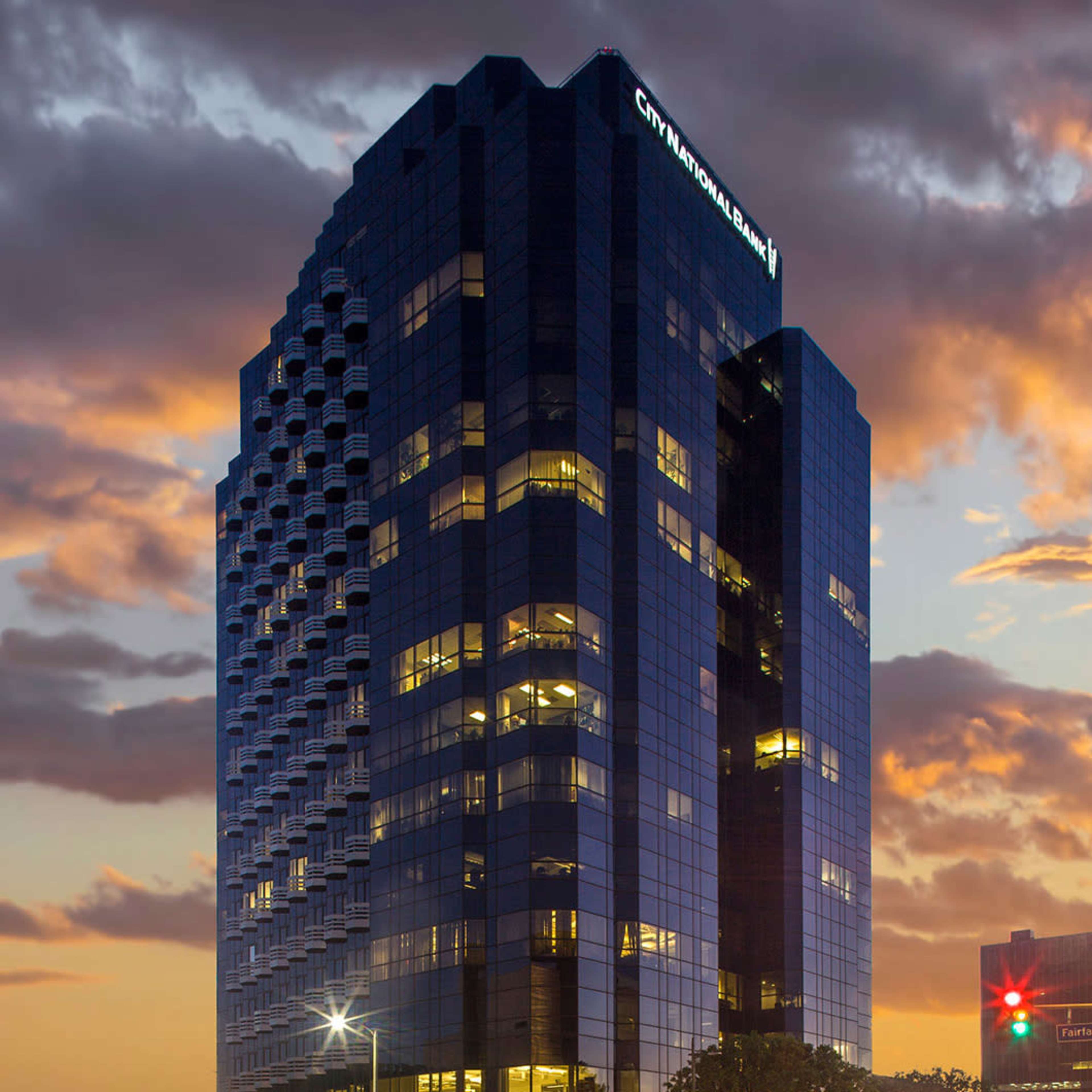 A tall, modern glass office building stands against a sunset sky, with the name "City National Bank" illuminated on its rooftop.