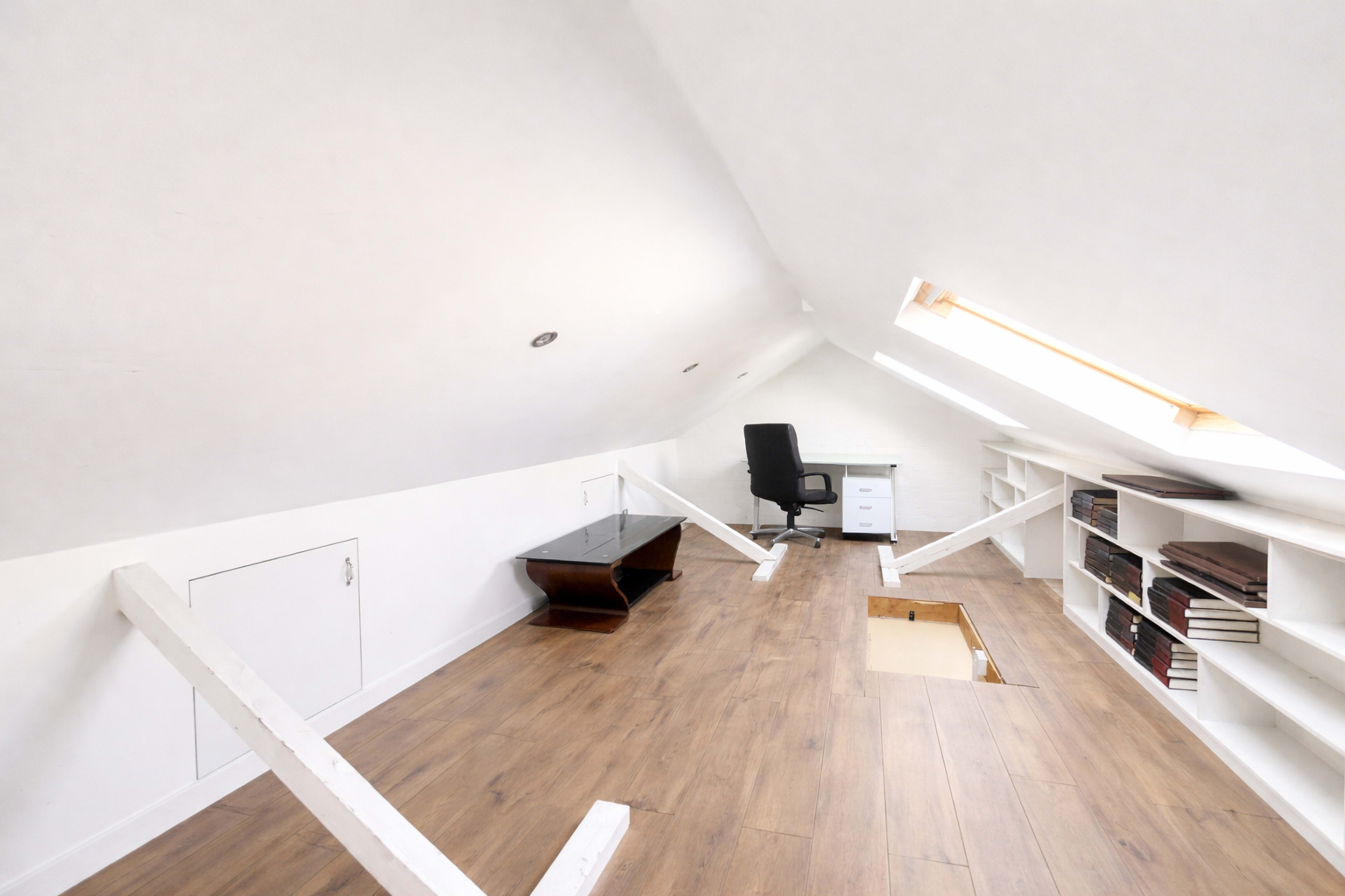 The image shows an empty attic space with a sloped ceiling, a black office chair, a small desk, and shelves filled with books.