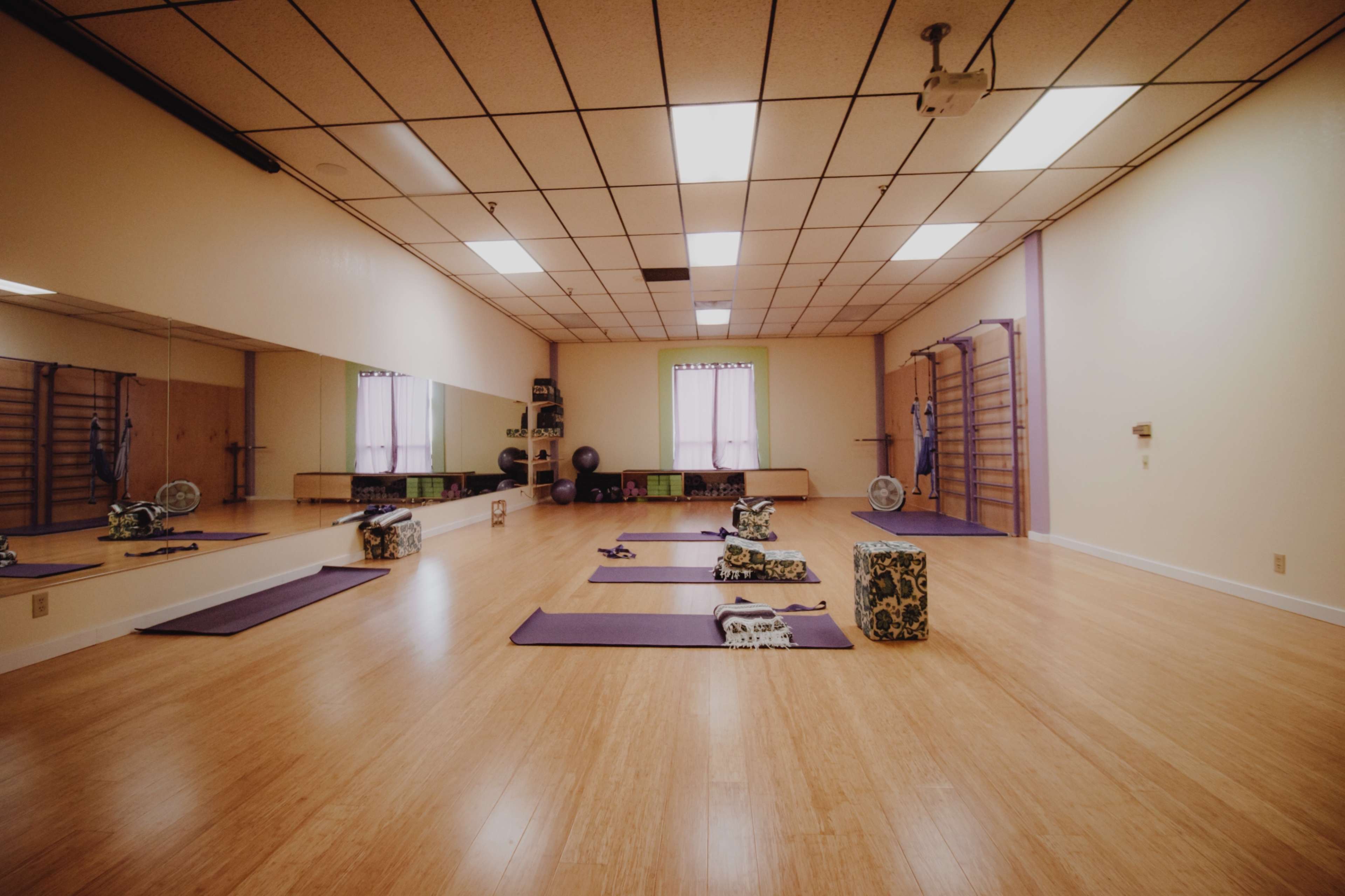 A spacious yoga studio with wooden flooring, yoga mats arranged on the floor, and equipment such as blocks and straps positioned around the room.