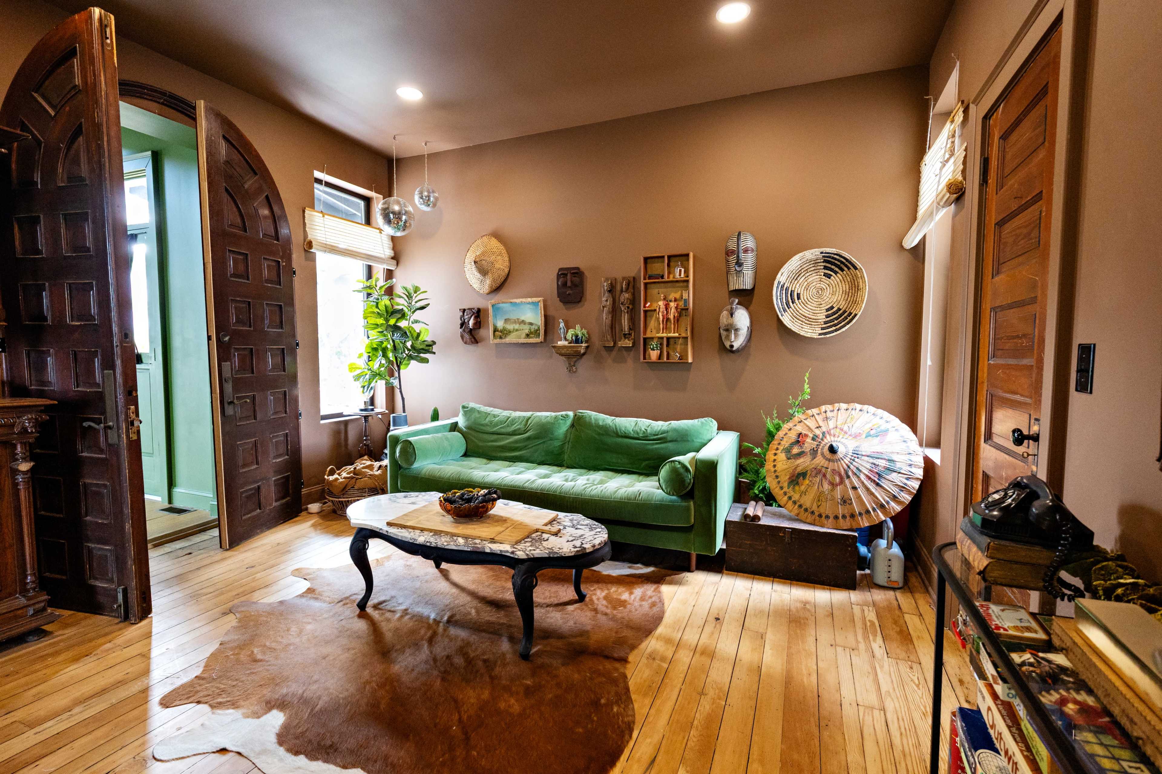The image shows a cozy living room with a green sofa, wooden floor, various wall decorations, and an open door leading to another room.