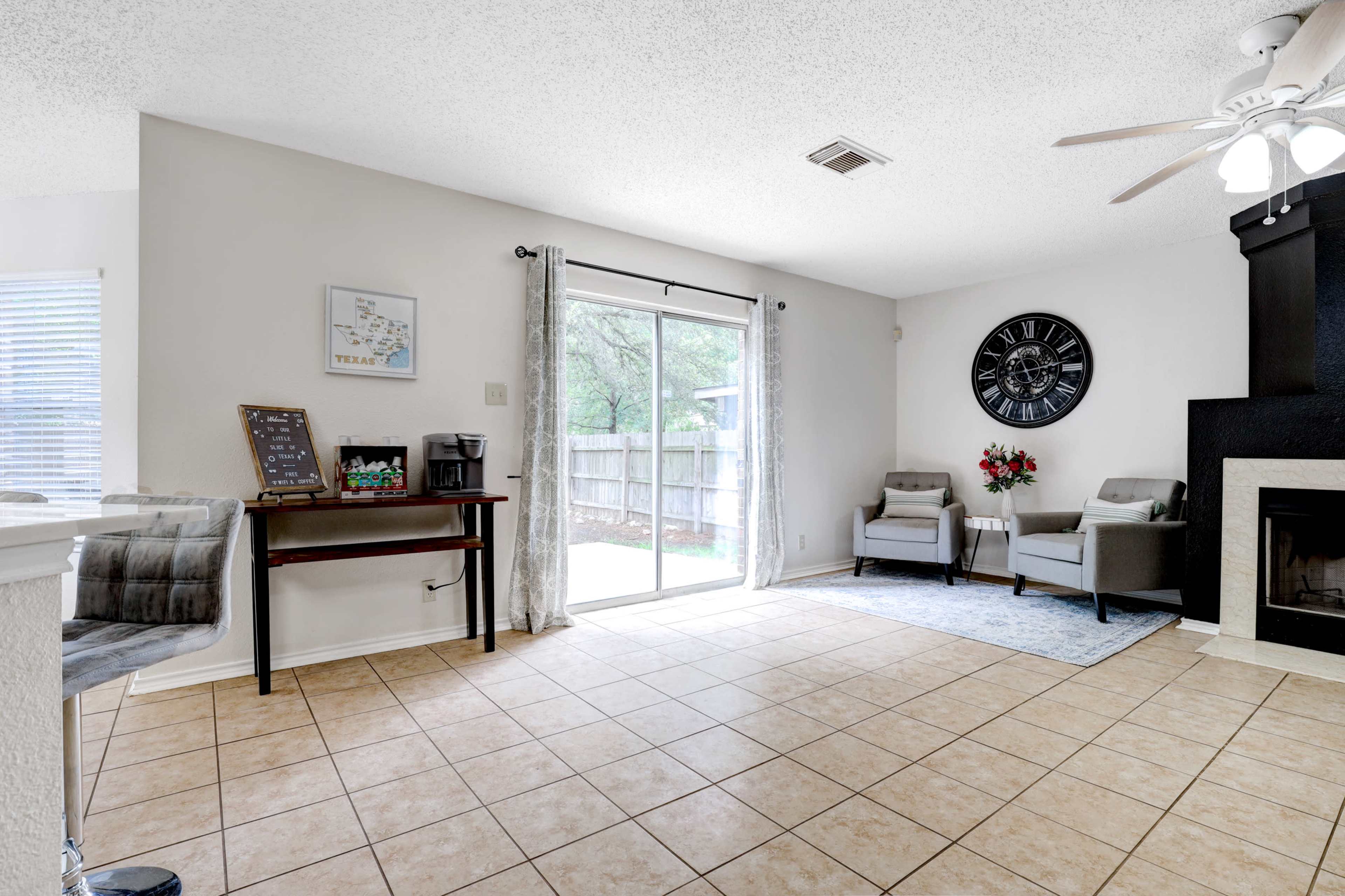 A living area with tiled floors, light walls, a sliding glass door, two armchairs, a decorative wall clock, and a corner fireplace.