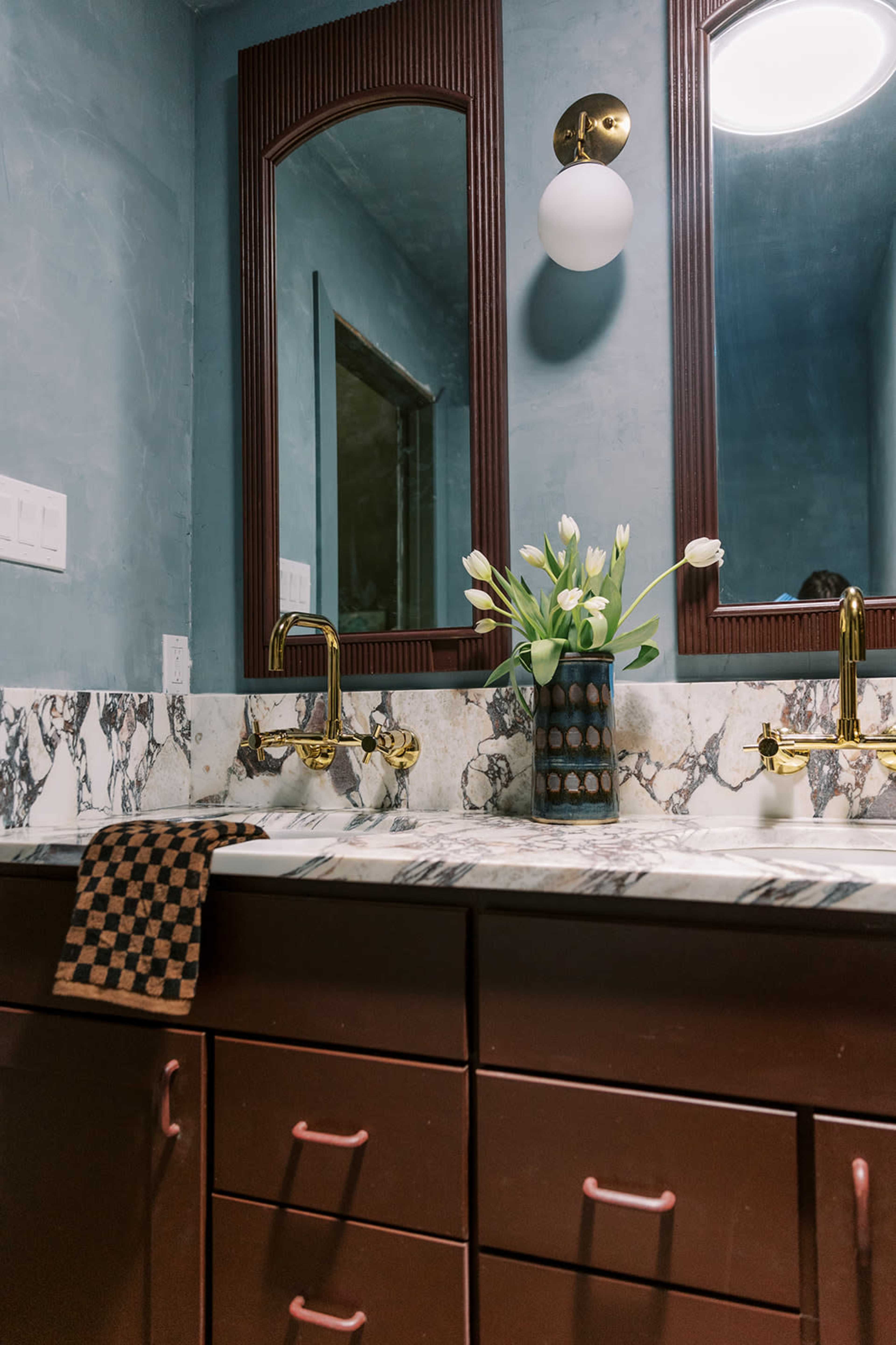 A bathroom features a marble countertop with two gold faucets, twin mirrors, and a vase of white tulips on a dark wooden vanity.