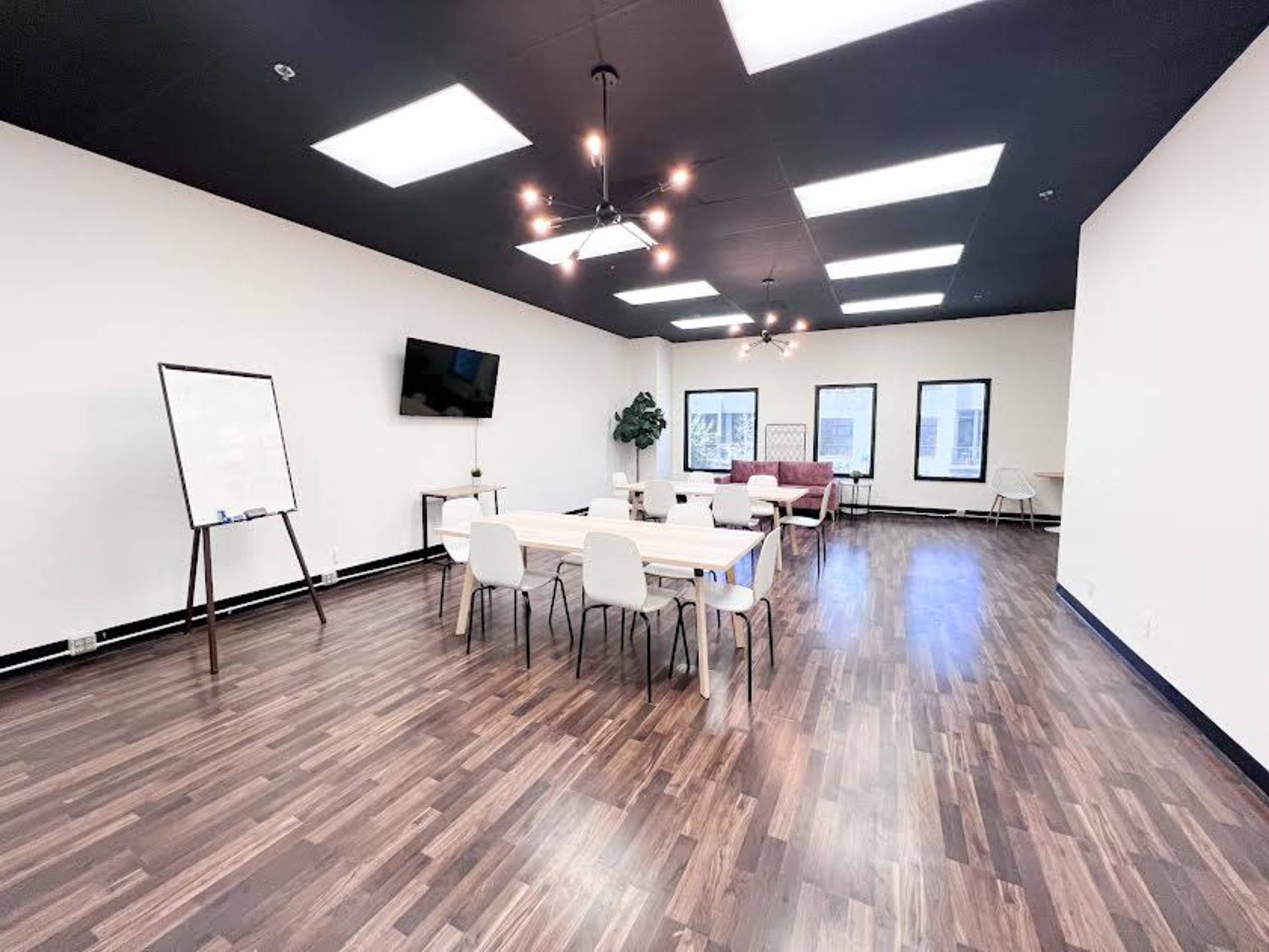 A modern, well-lit meeting room with a large table, several chairs, a whiteboard, a television mounted on the wall, and a couch.
