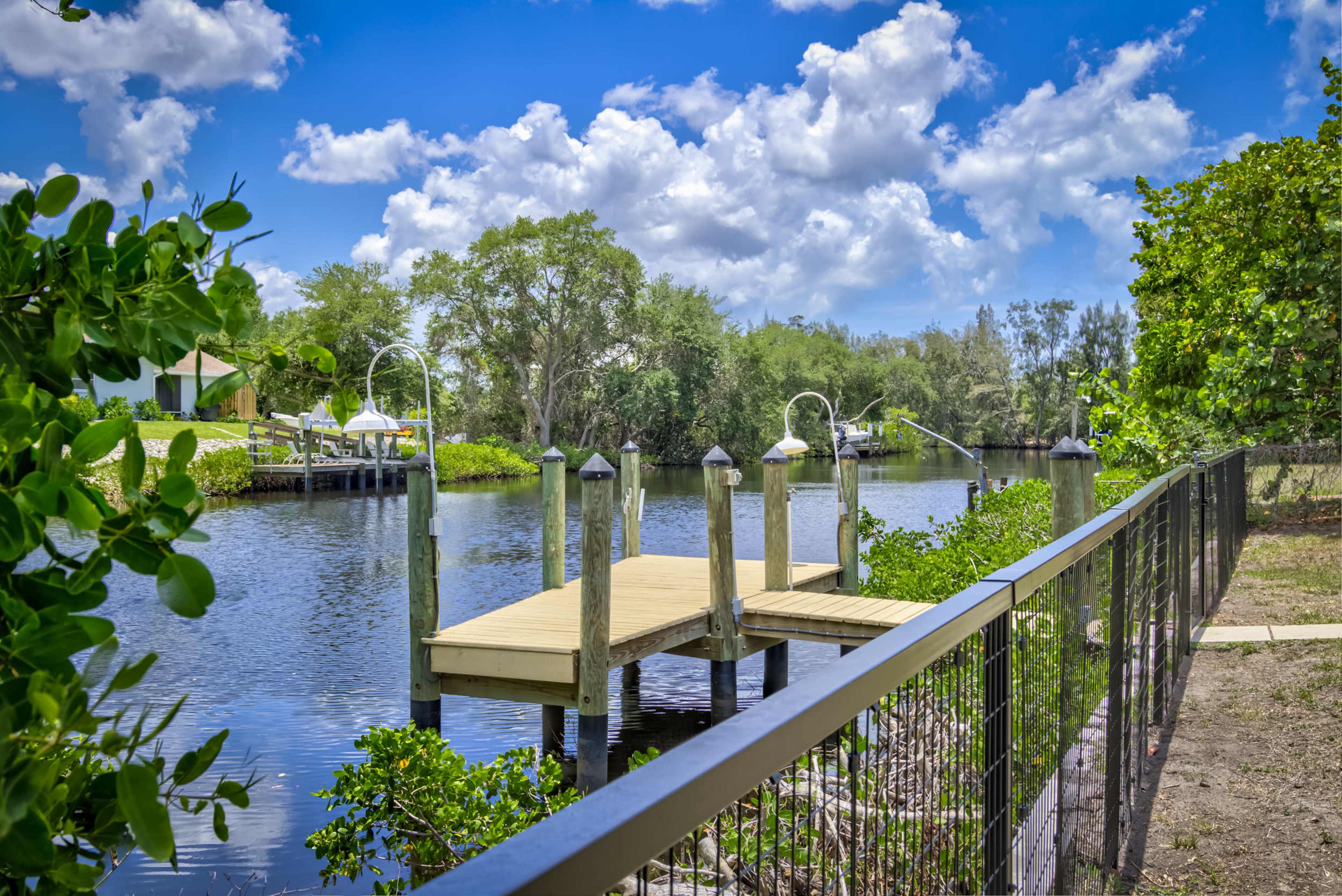 A wooden dock with two lamps extends over a calm waterway, bordered by greenery and a fence on one side.