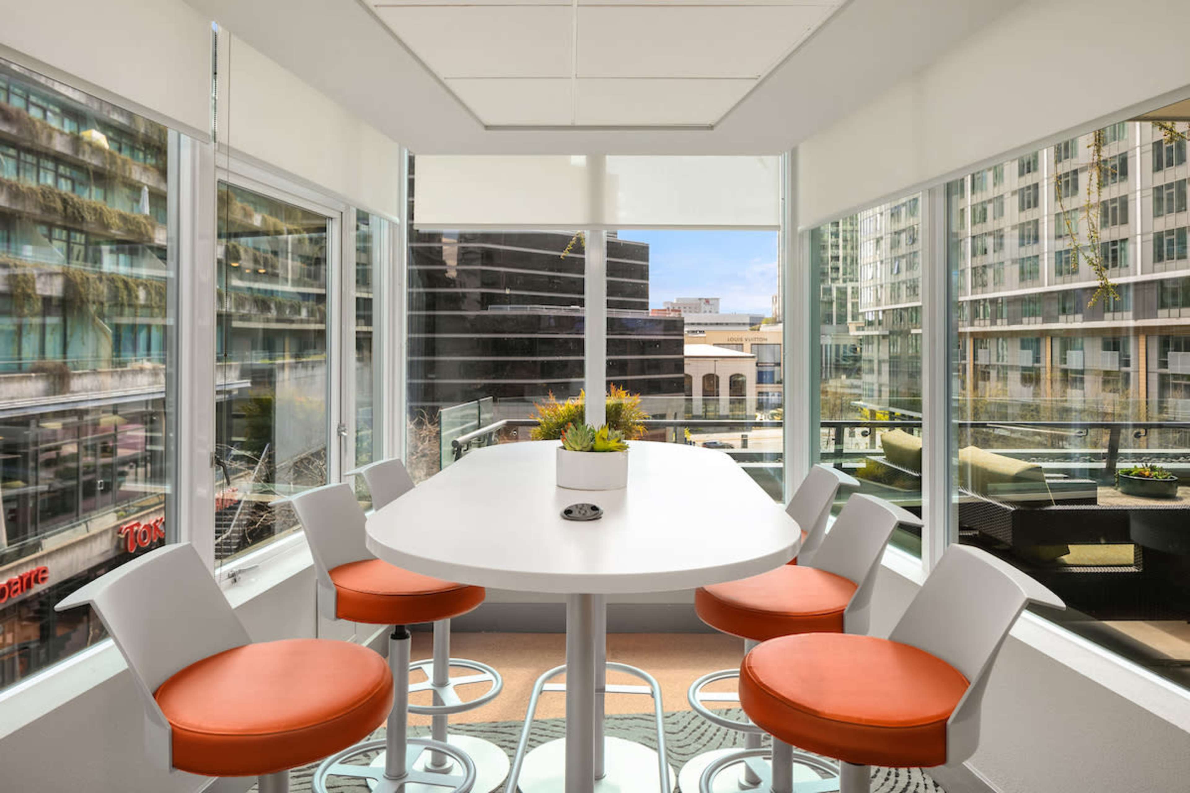 A bright, modern meeting room features a round white table surrounded by orange-accented chairs, with large windows showcasing a cityscape.