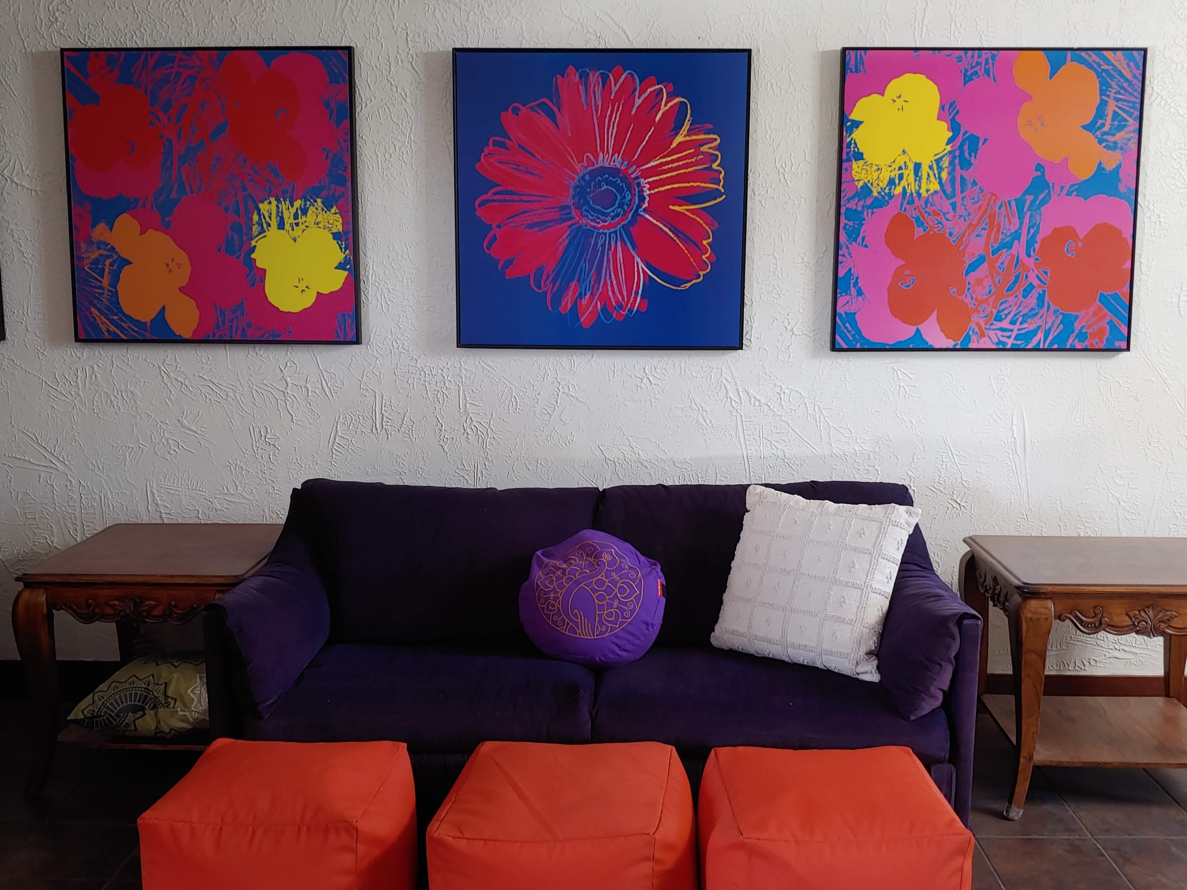 The image shows a purple sofa with a colorful throw pillow in front of three vibrant floral artworks hanging on the wall above it, complemented by orange ottomans.