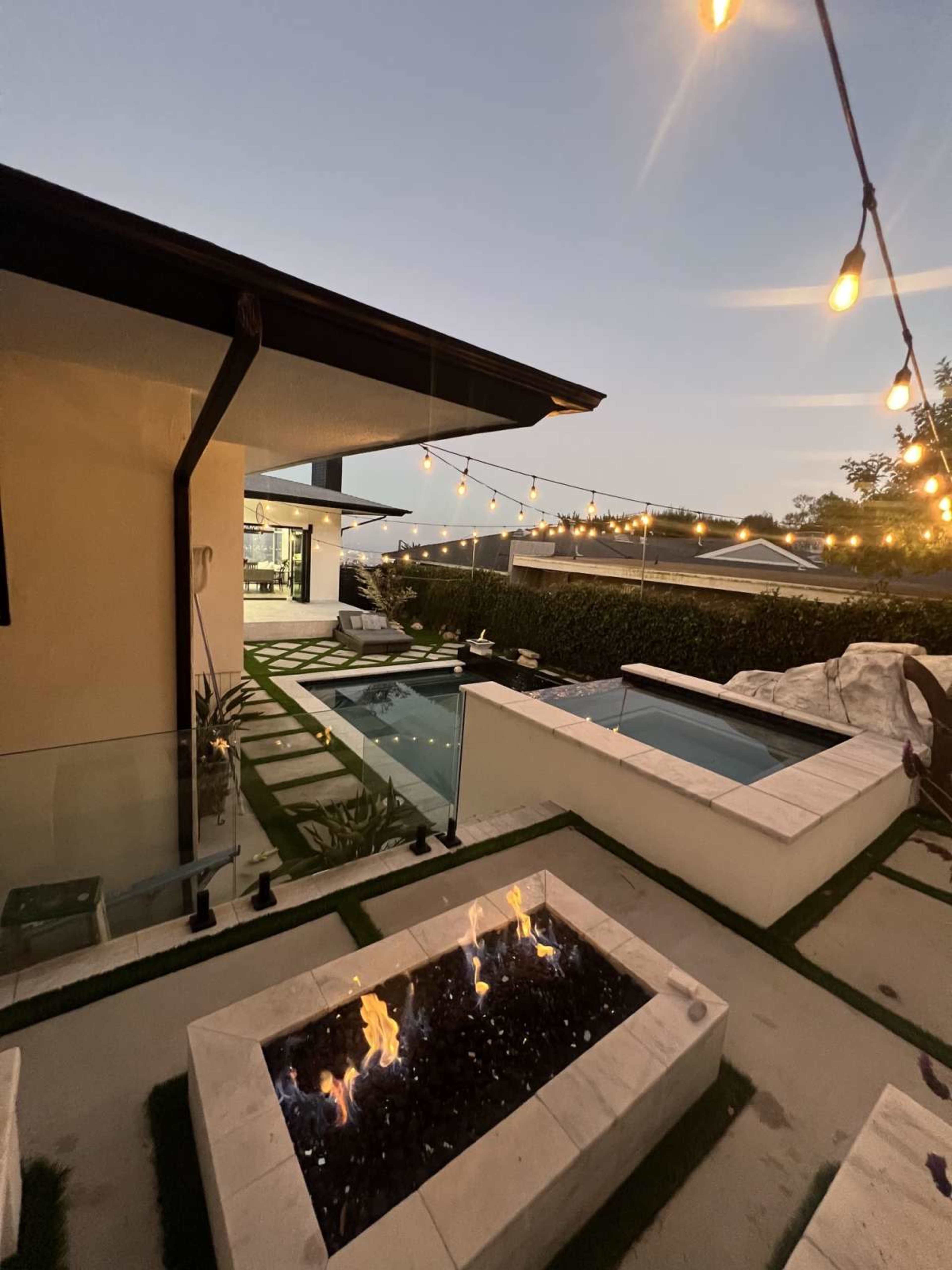 The image shows a modern outdoor space featuring a fire pit, a small pool, and string lights, with lush landscaping and a contemporary building in the background.