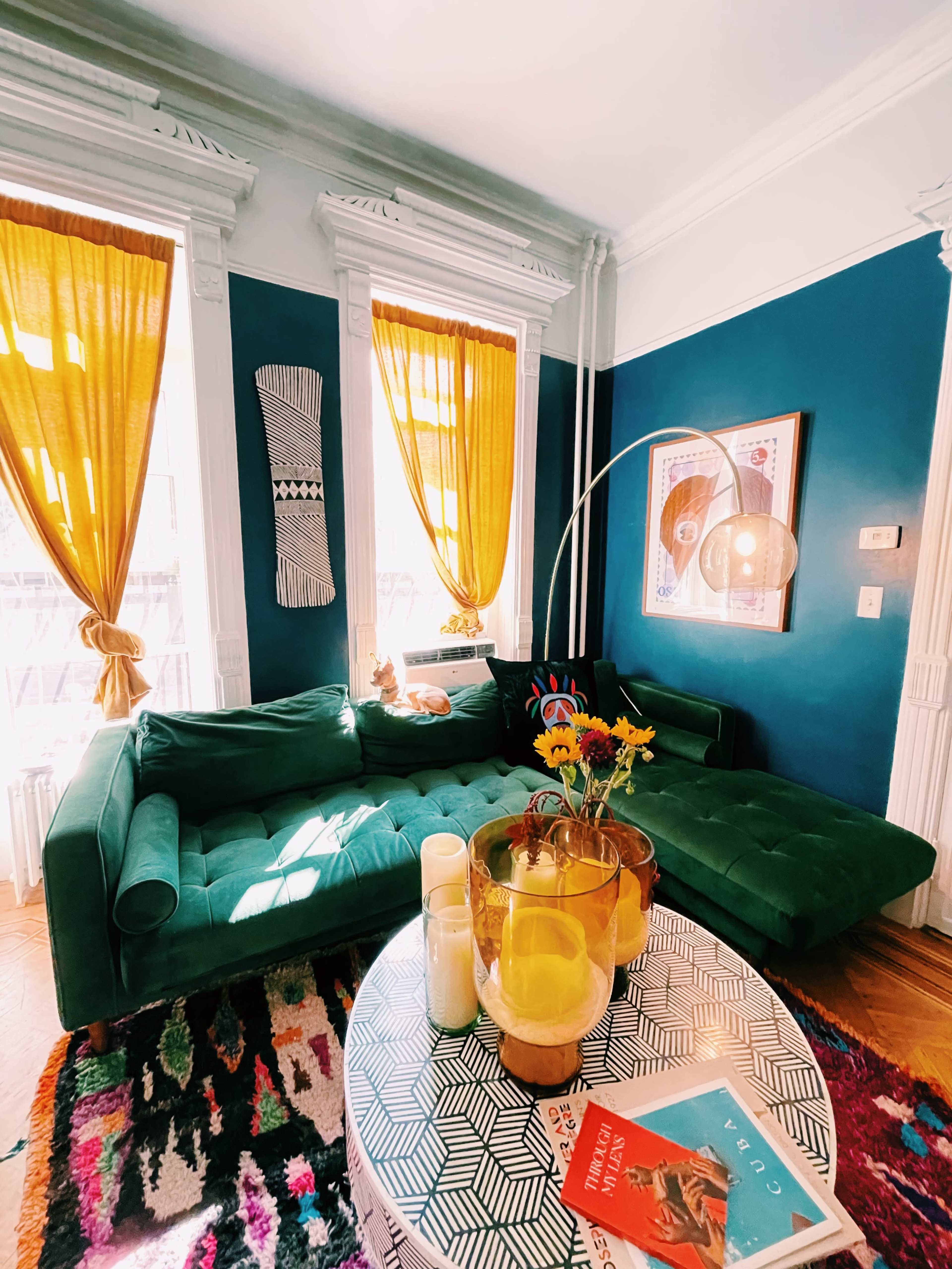 A colorful living room with a green sectional sofa, a round table with books and a vase of flowers, and large windows with yellow curtains.