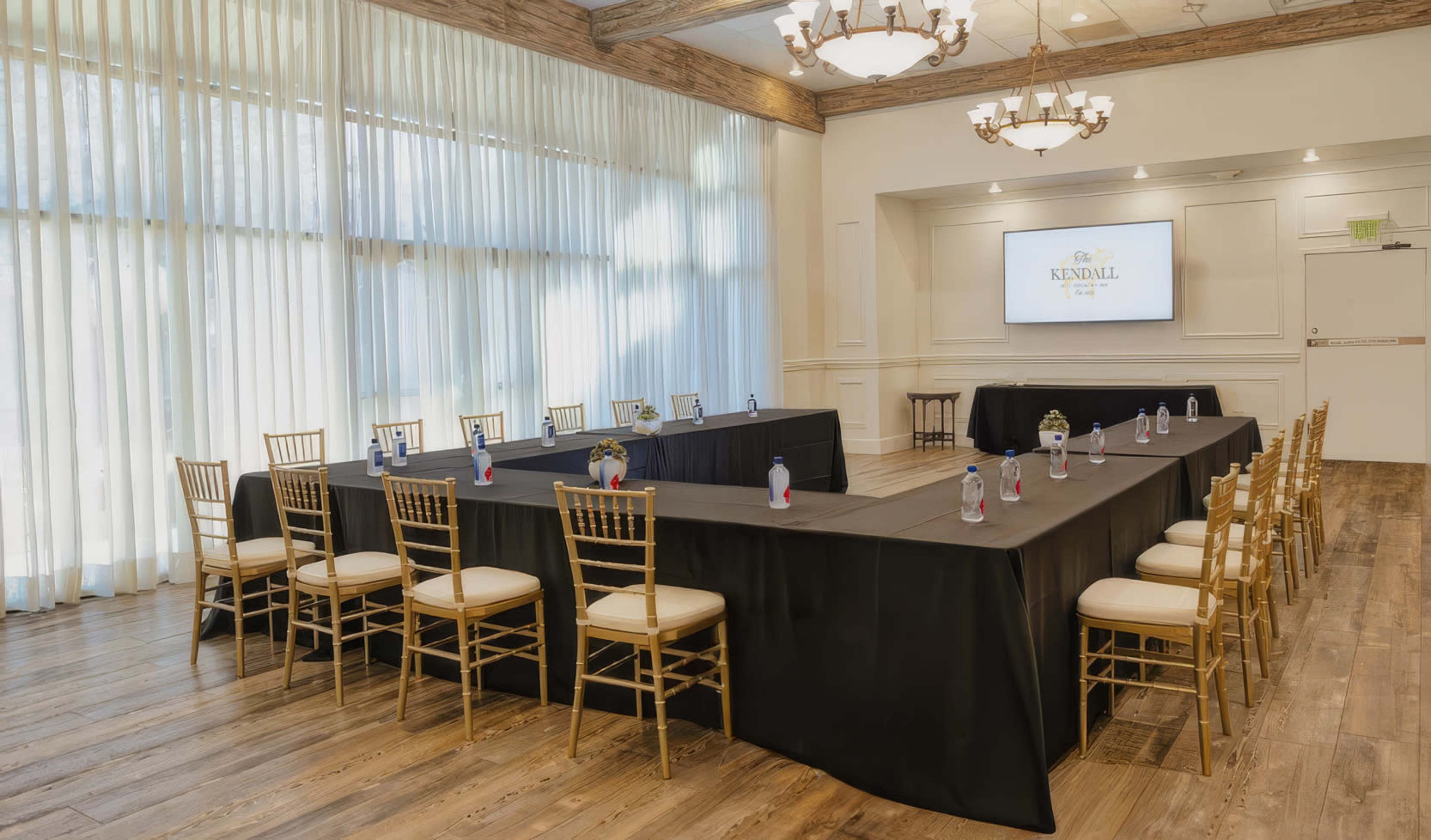 A conference room features a large rectangular setup of black tables surrounded by gold chairs, with natural light streaming in through tall windows.