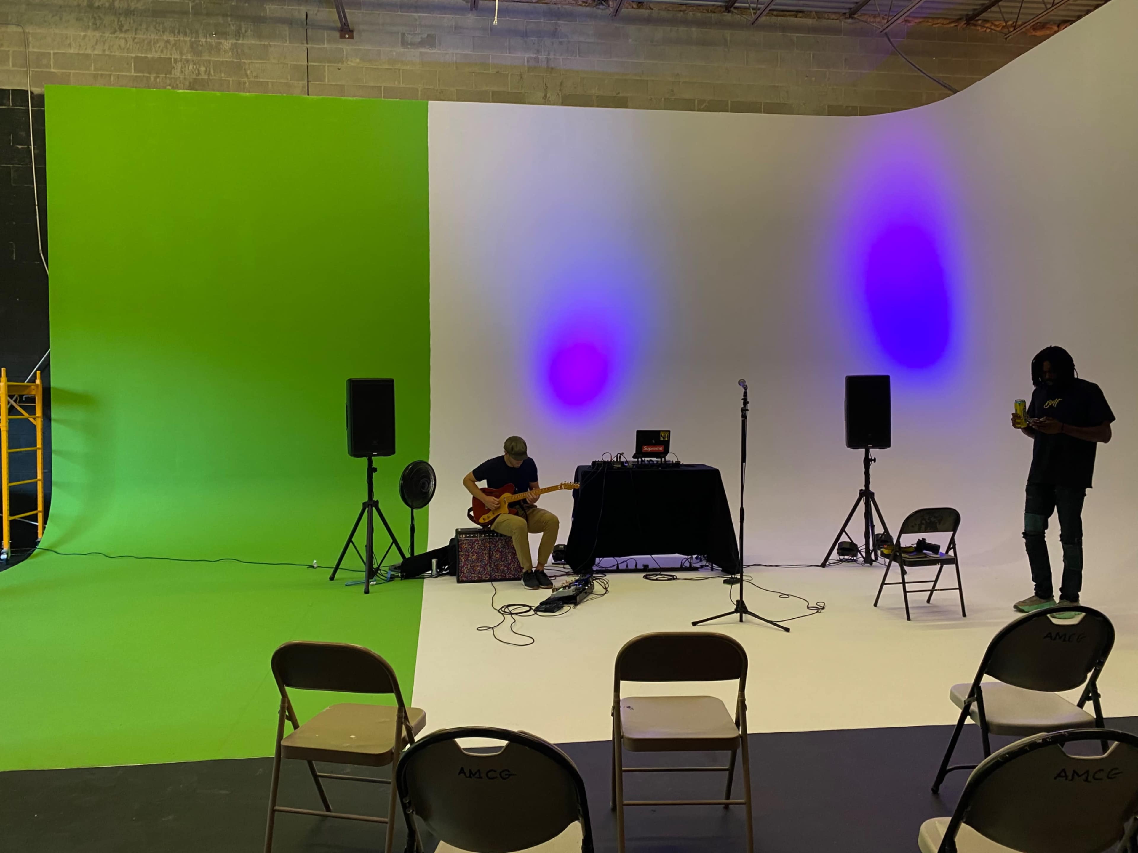 A musician plays guitar on stage with a sound setup in front of a backdrop that is divided into green and white sections, with speakers and chairs arranged for an audience.