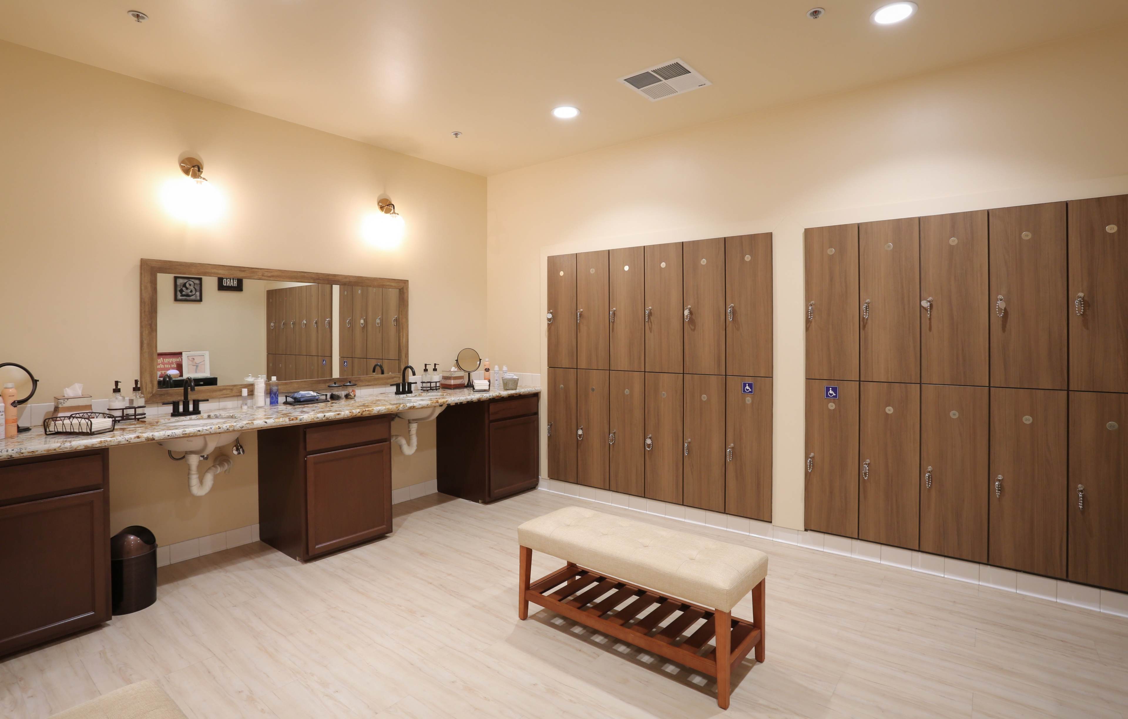 The image shows a well-lit locker room featuring a row of wooden lockers, a countertop with sinks, and a bench in the center.
