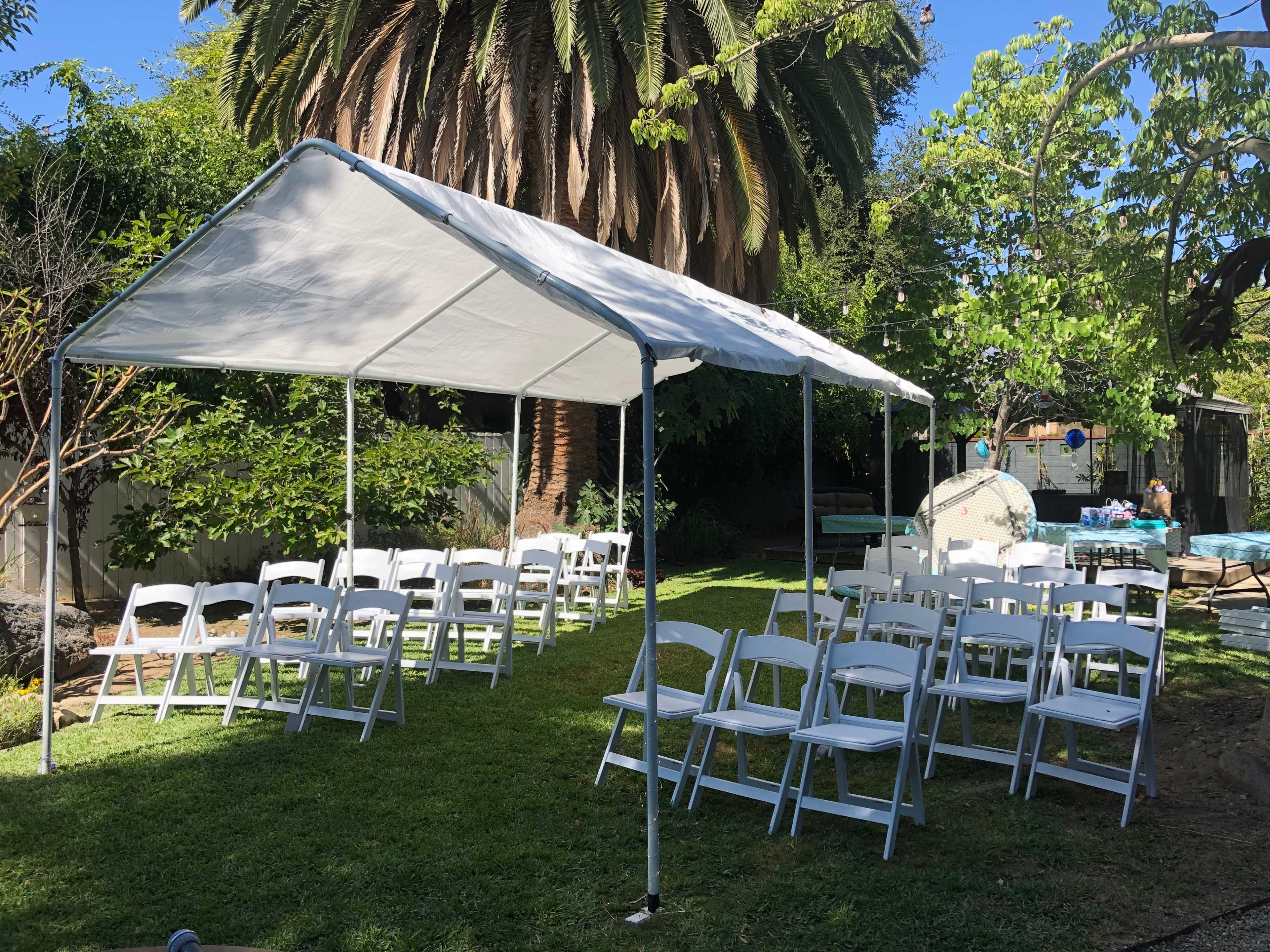 A white tent is set up on grass with rows of white chairs arranged underneath it, surrounded by trees and greenery.