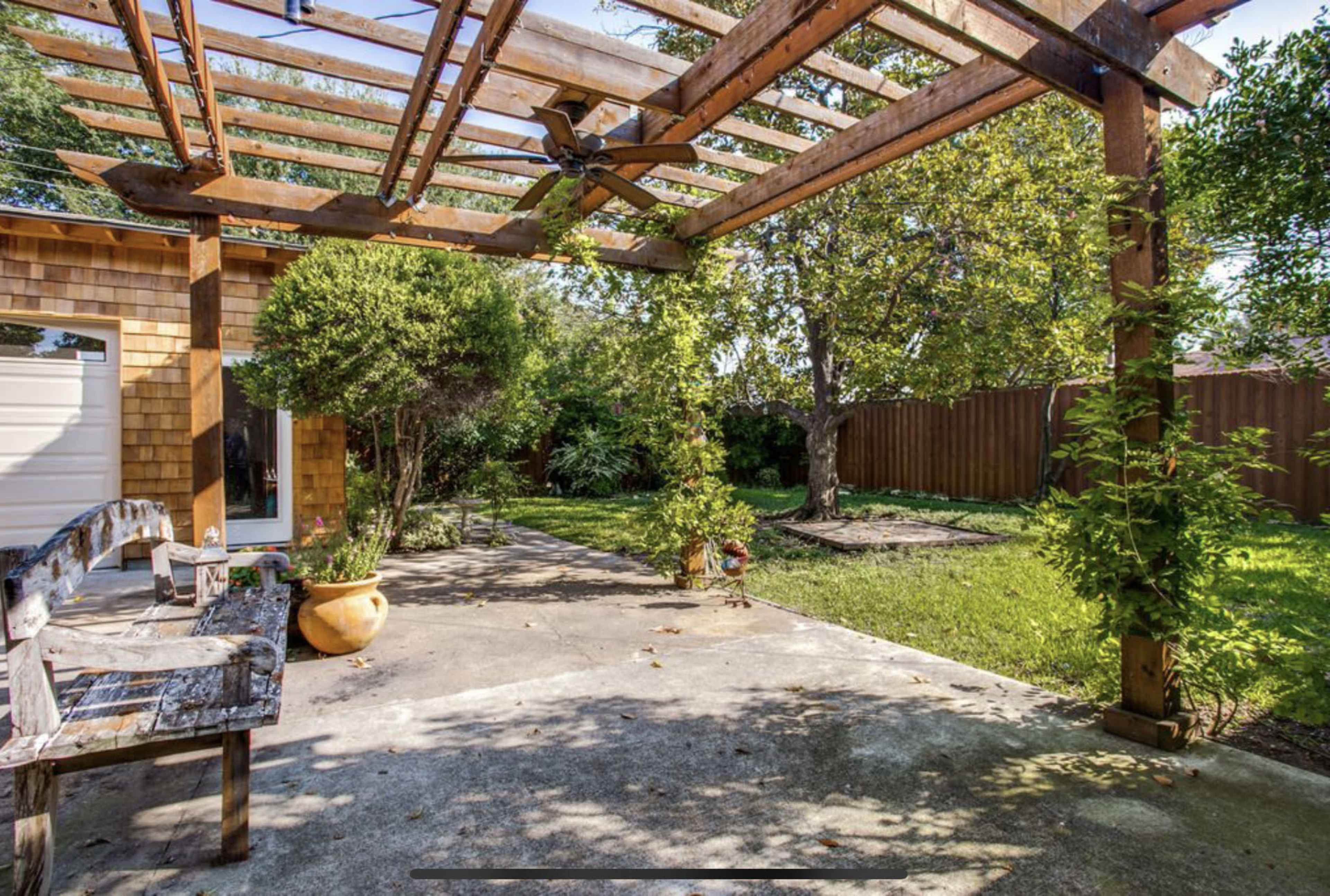 An outdoor space features a wooden pergola with climbing plants, a stone pathway, and greenery in the background.