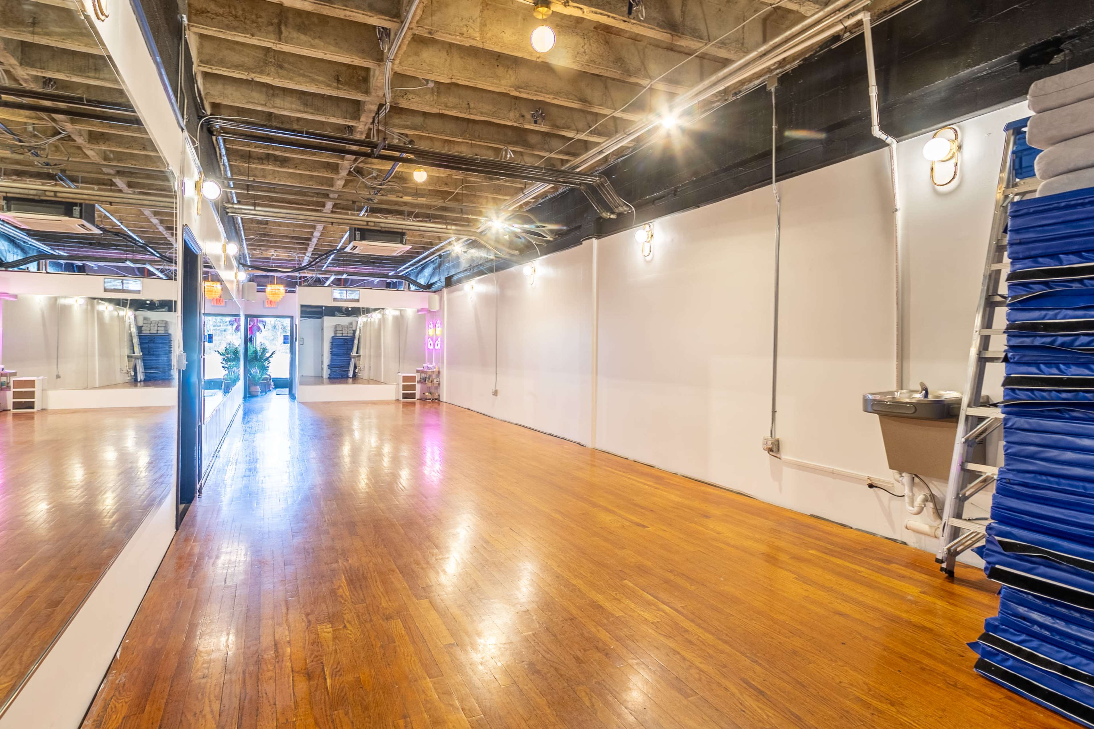 A spacious indoor room with polished wooden floors, large mirrors on one wall, and fitness mats stacked against another wall.
