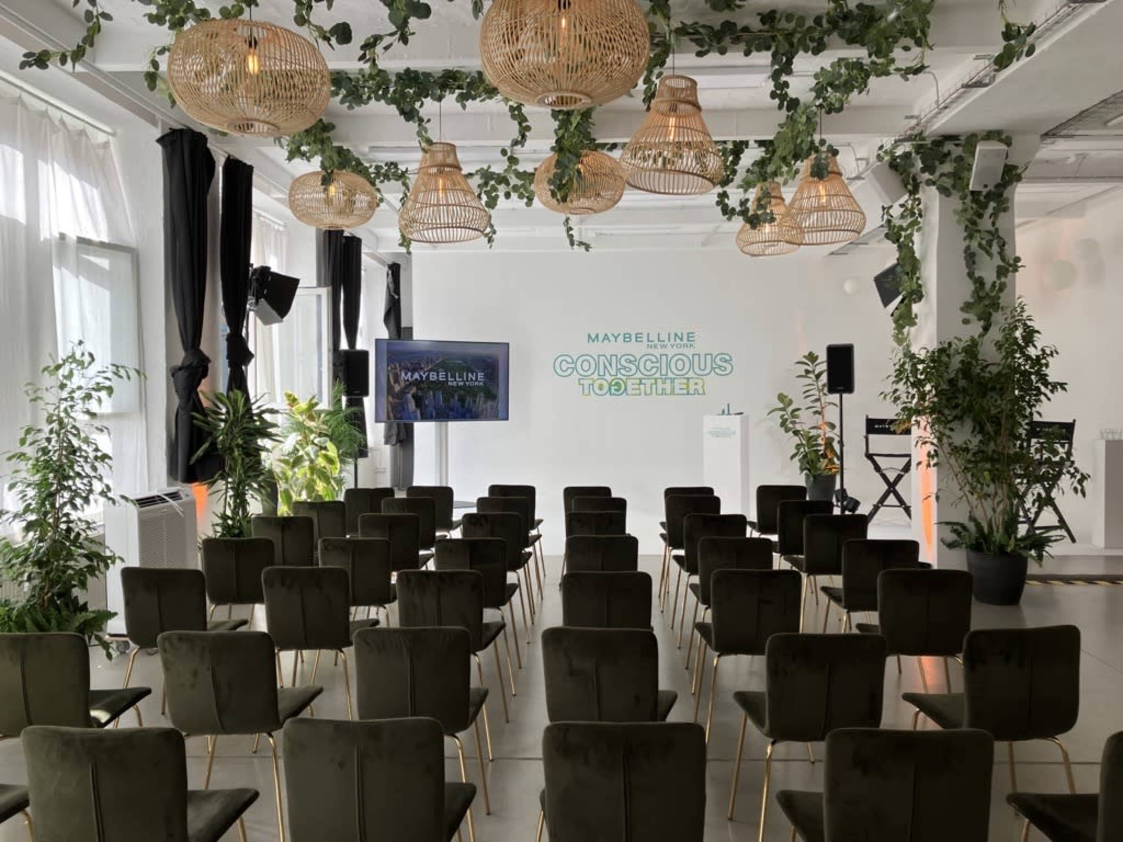 A modern event space set up with rows of green chairs facing a stage adorned with plants, hanging lights, and a large screen displaying the Maybelline logo and the phrase "CONSCIOUS TOGETHER."