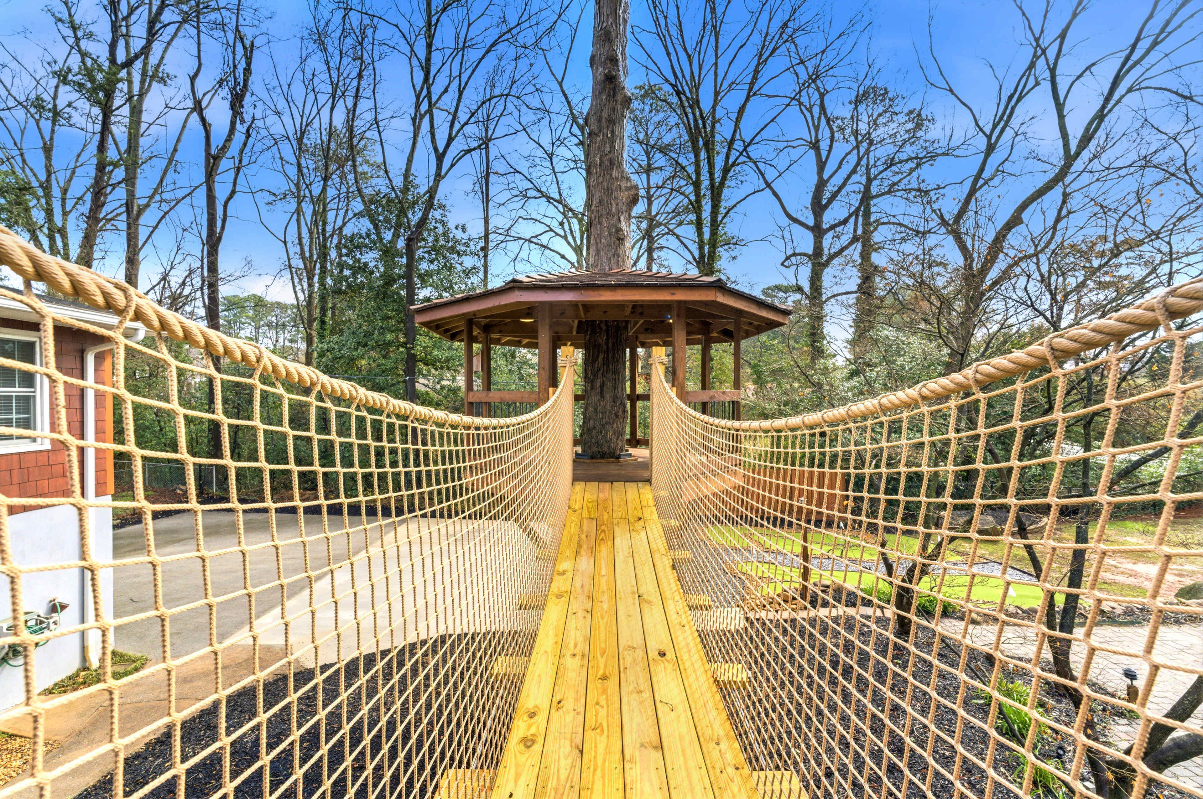 A wooden suspension bridge with rope handrails leads to a gazebo surrounded by bare trees.