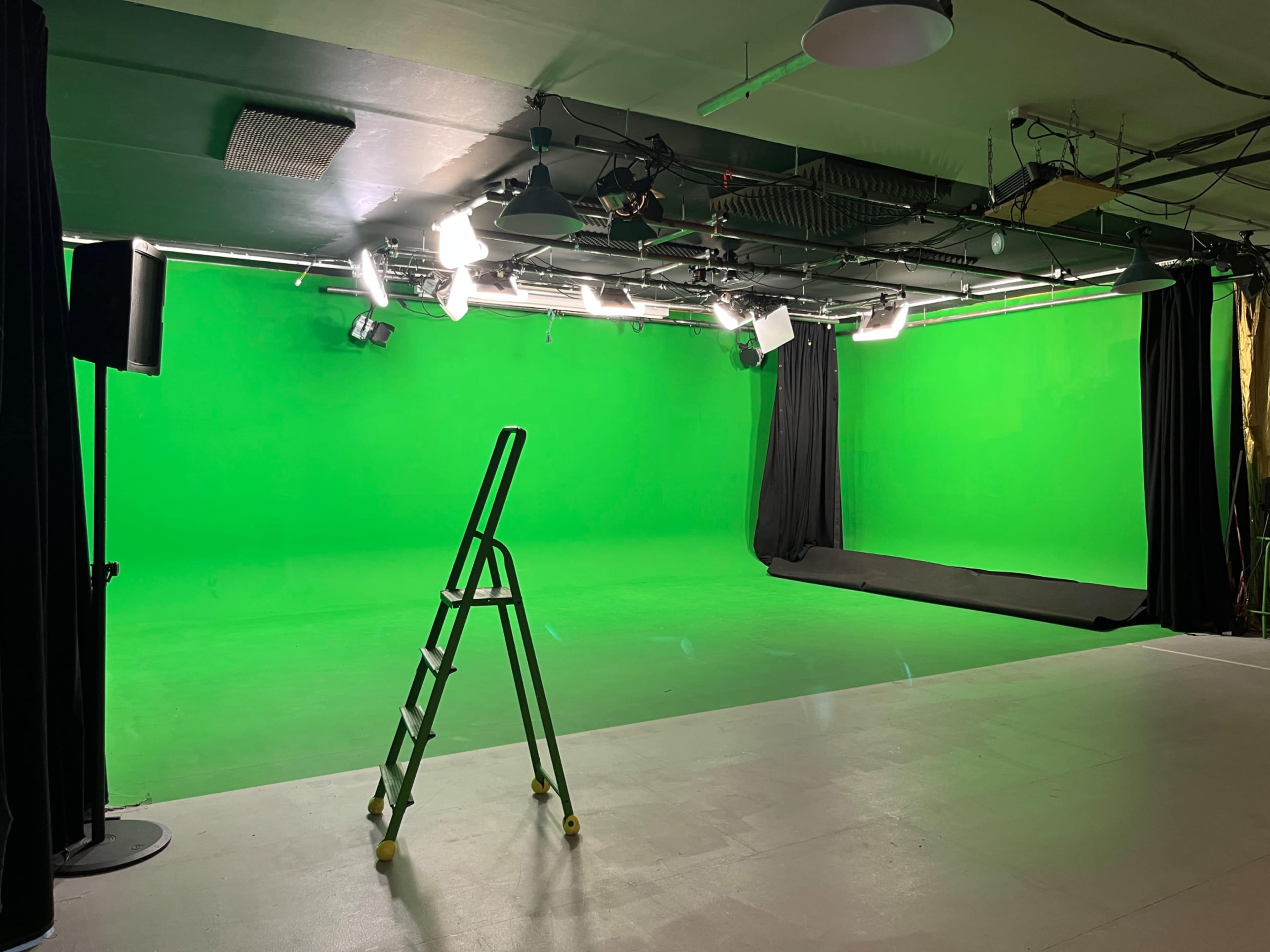 The image shows an empty green screen studio with a stepladder in the foreground and soft lighting overhead.