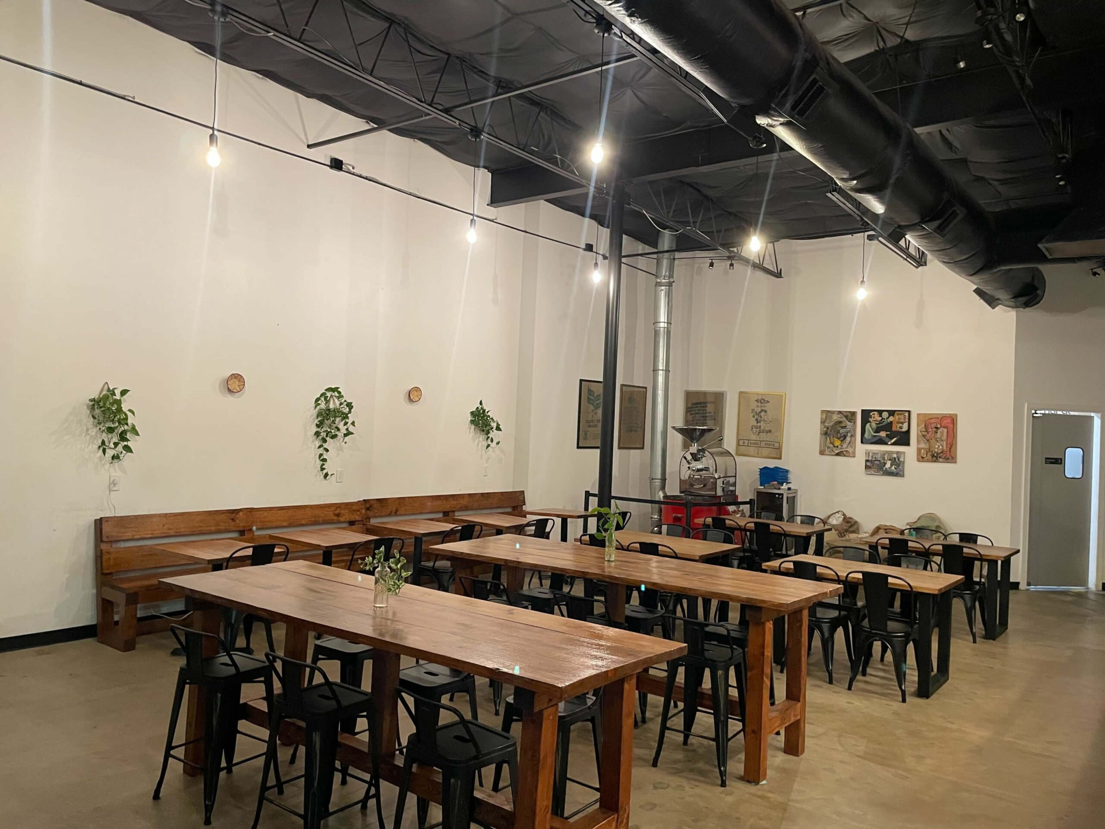 The image shows a spacious, industrial-style café featuring wooden tables and chairs, with plants and artwork adorning the white walls.