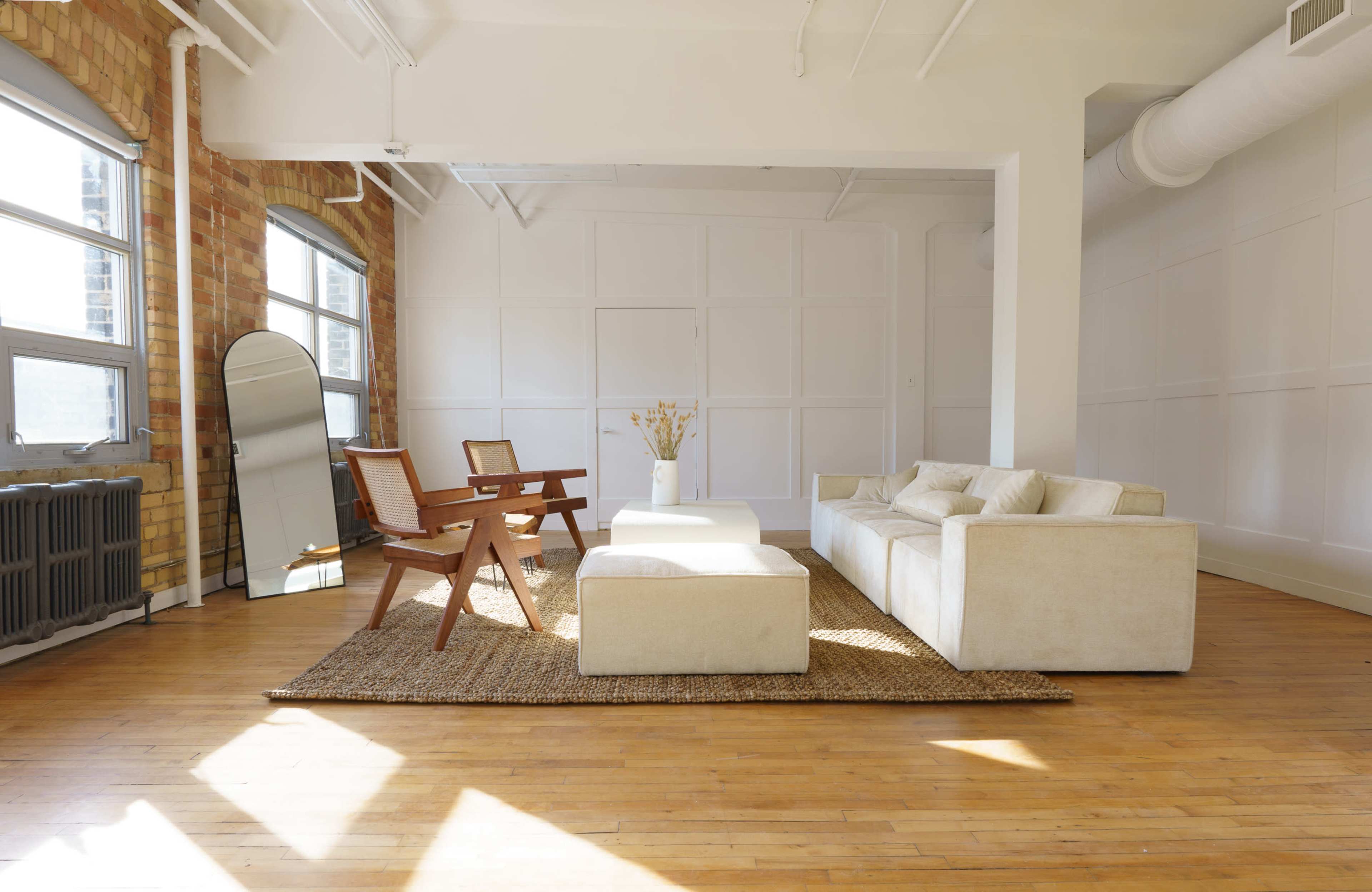 A bright and spacious living room with a beige sofa, a matching coffee table, two wooden chairs, a mirror, and a large area rug on a hardwood floor.