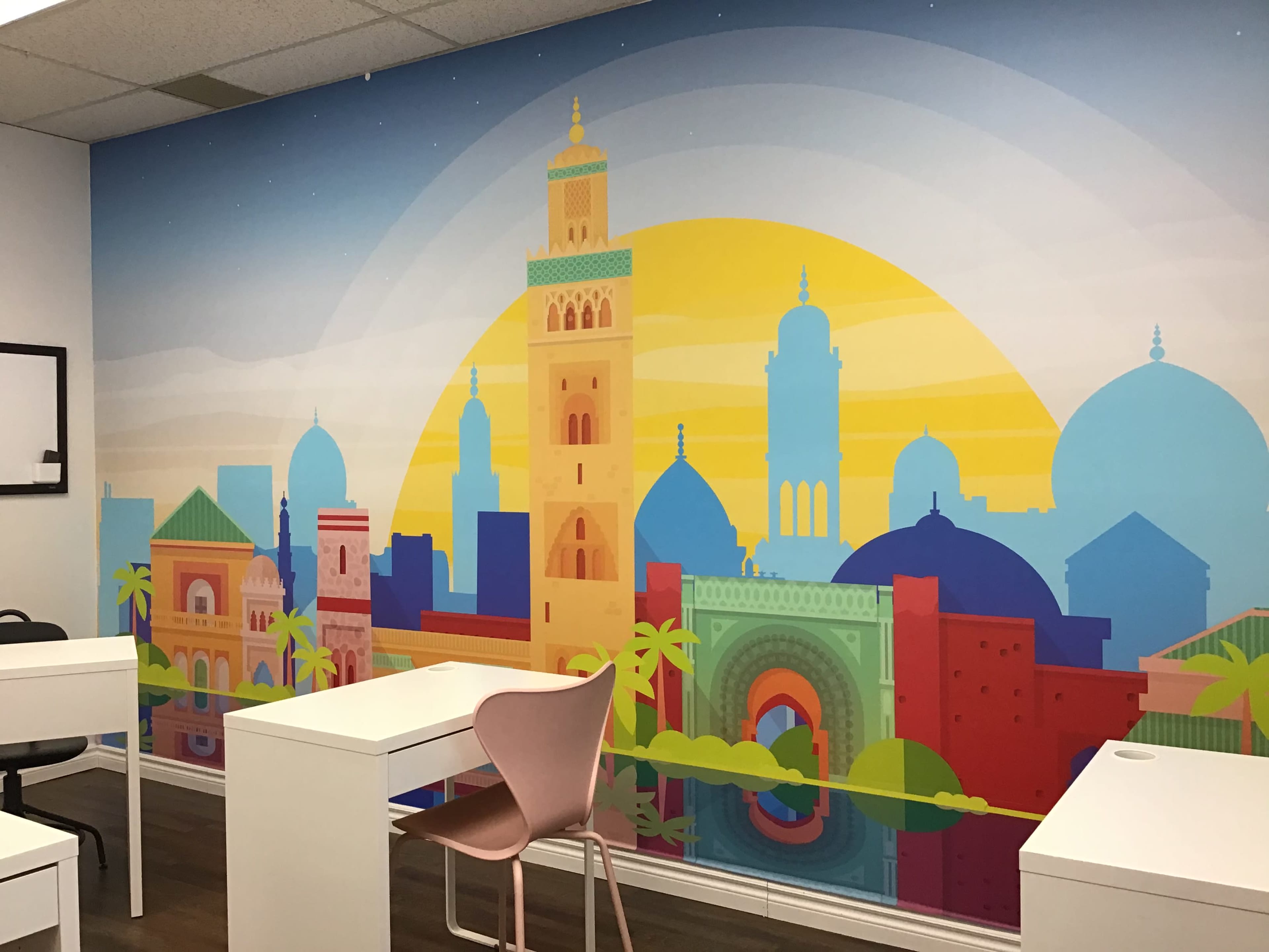 A brightly colored mural featuring a skyline with various architectural styles, set against a large yellow sun, in a room with white desks and a pink chair.