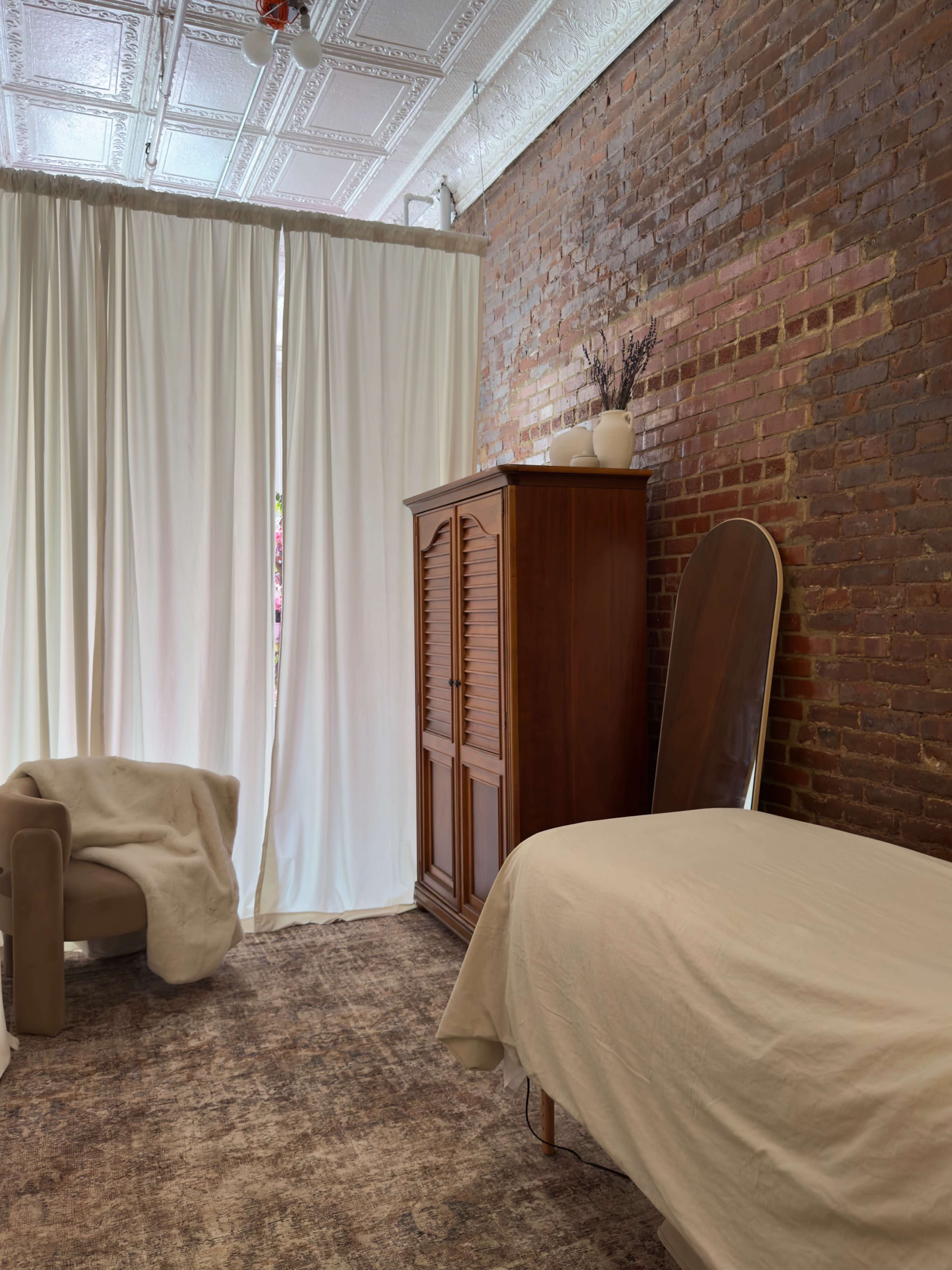 The image shows a room with exposed brick walls, a bed covered with a light-colored blanket, a wooden wardrobe, and a cozy chair near sheer white curtains.