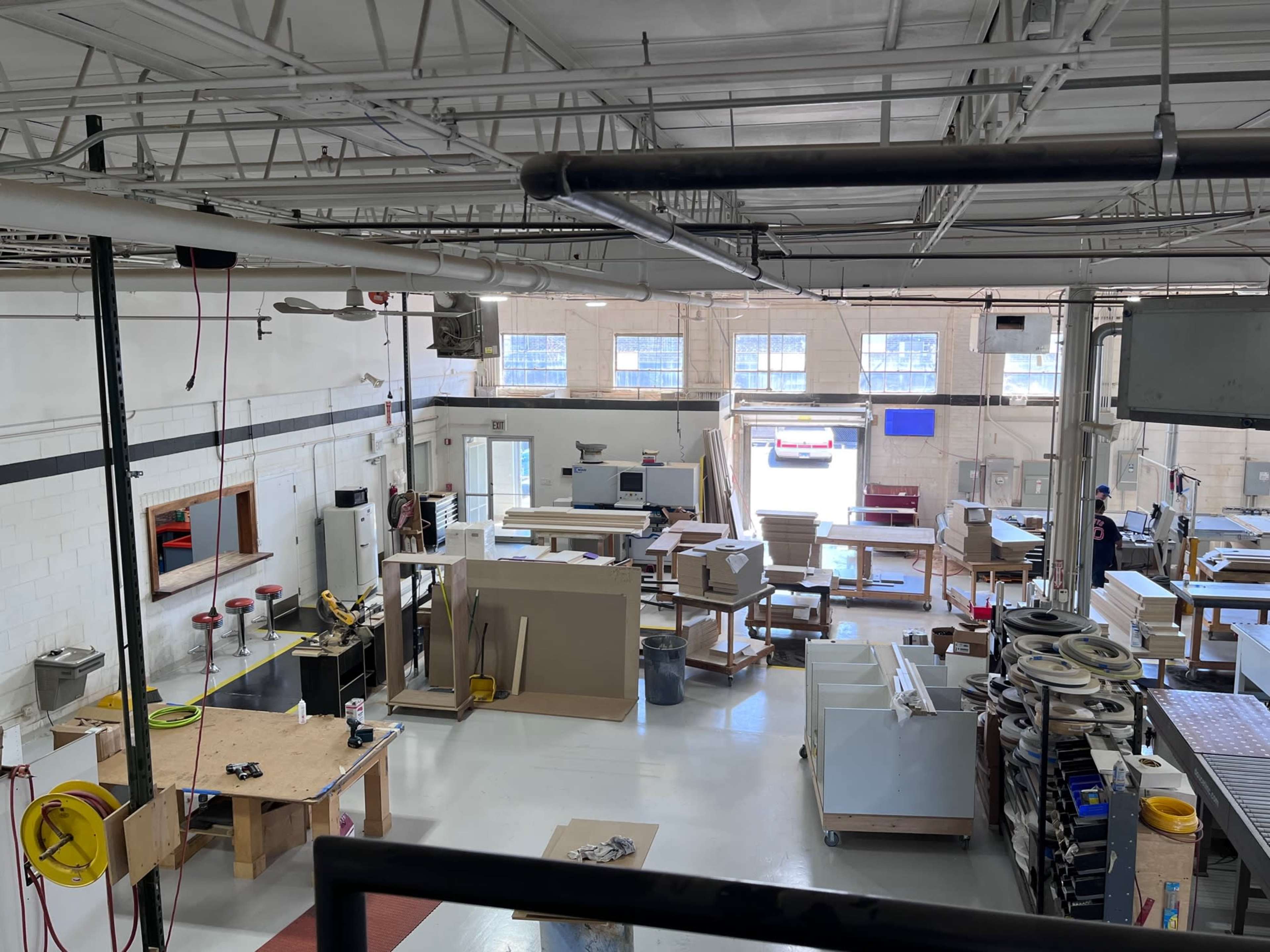 A large workshop filled with various woodworking tables, tools, and materials, with sunlight streaming through the windows.