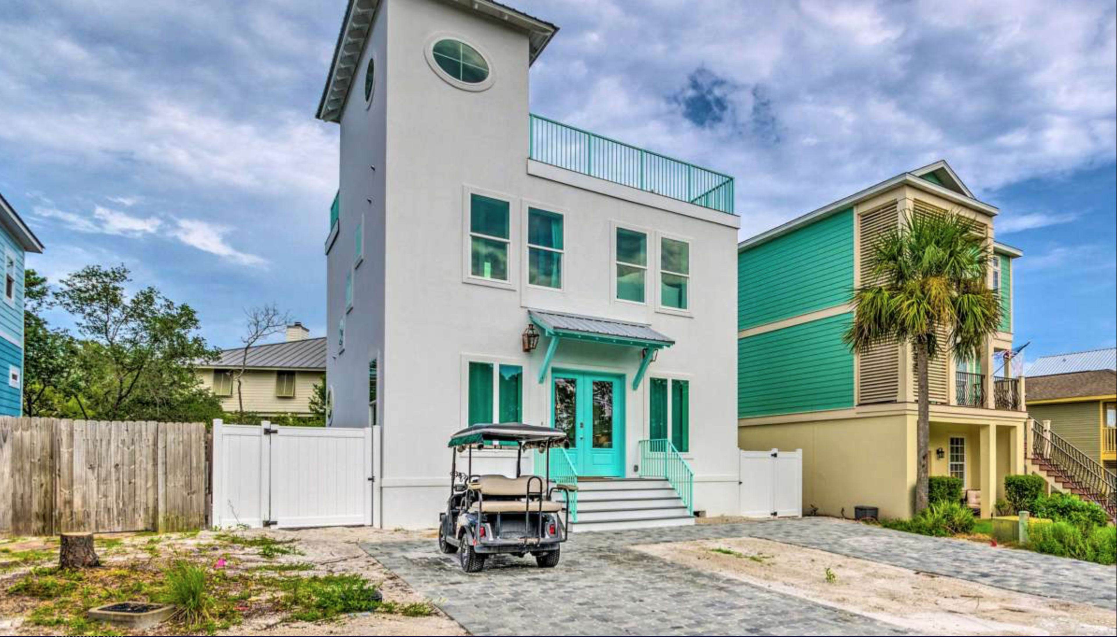 A modern three-story house with teal accents and a golf cart parked in front is surrounded by a landscaped yard and a wooden fence.