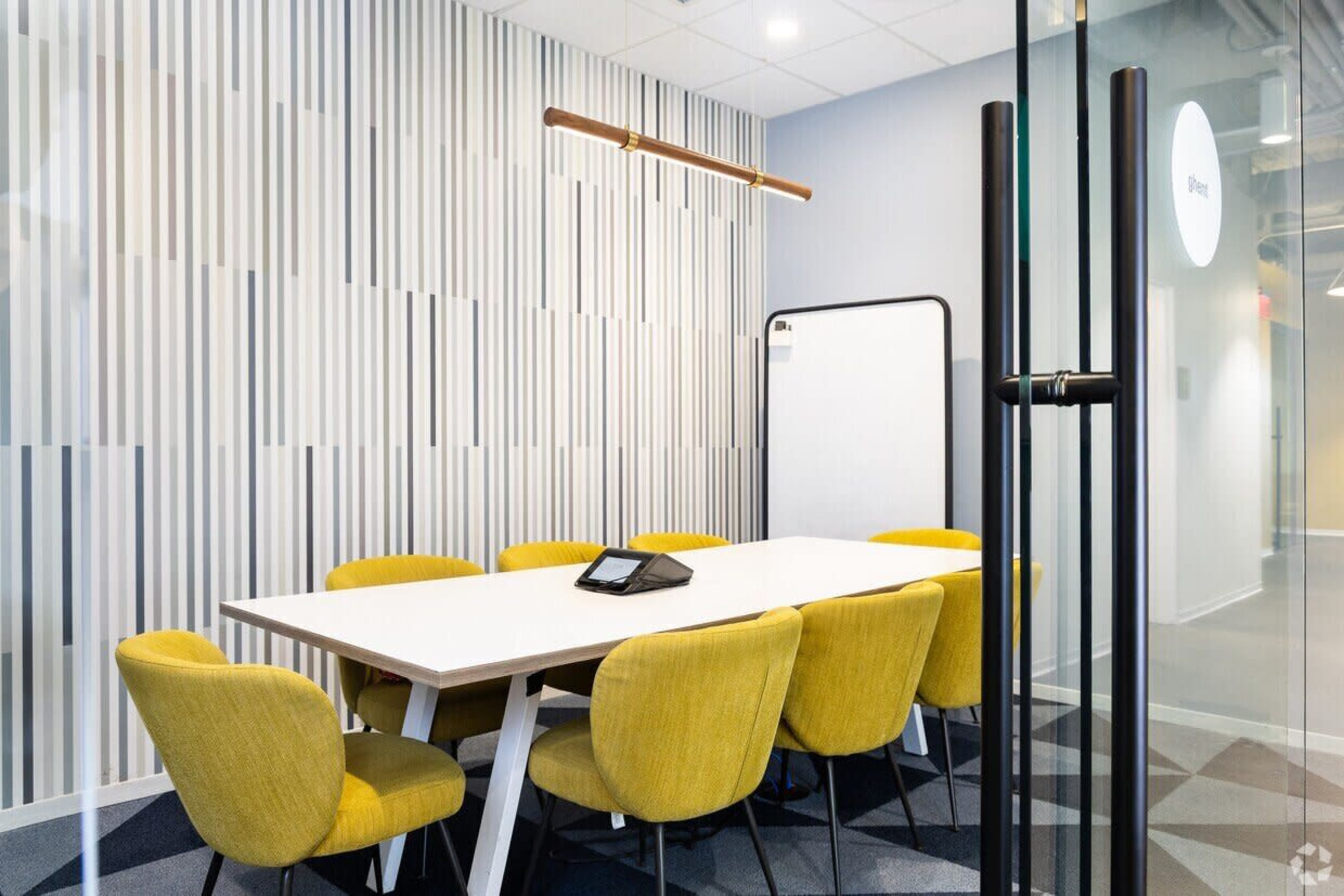 A modern conference room features a long white table surrounded by six yellow chairs, with a glass wall and a whiteboard nearby.