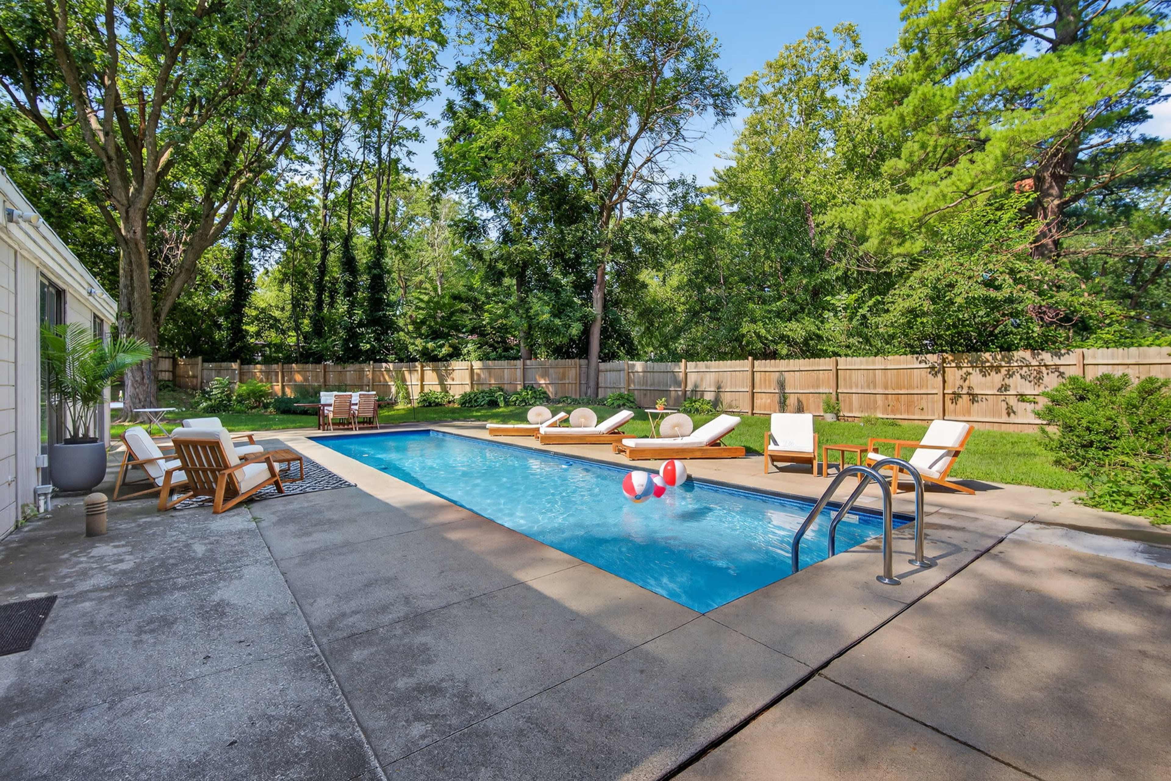 The image shows a rectangular swimming pool surrounded by lounge chairs and a wooden deck, set in a backyard with trees and a wooden fence.