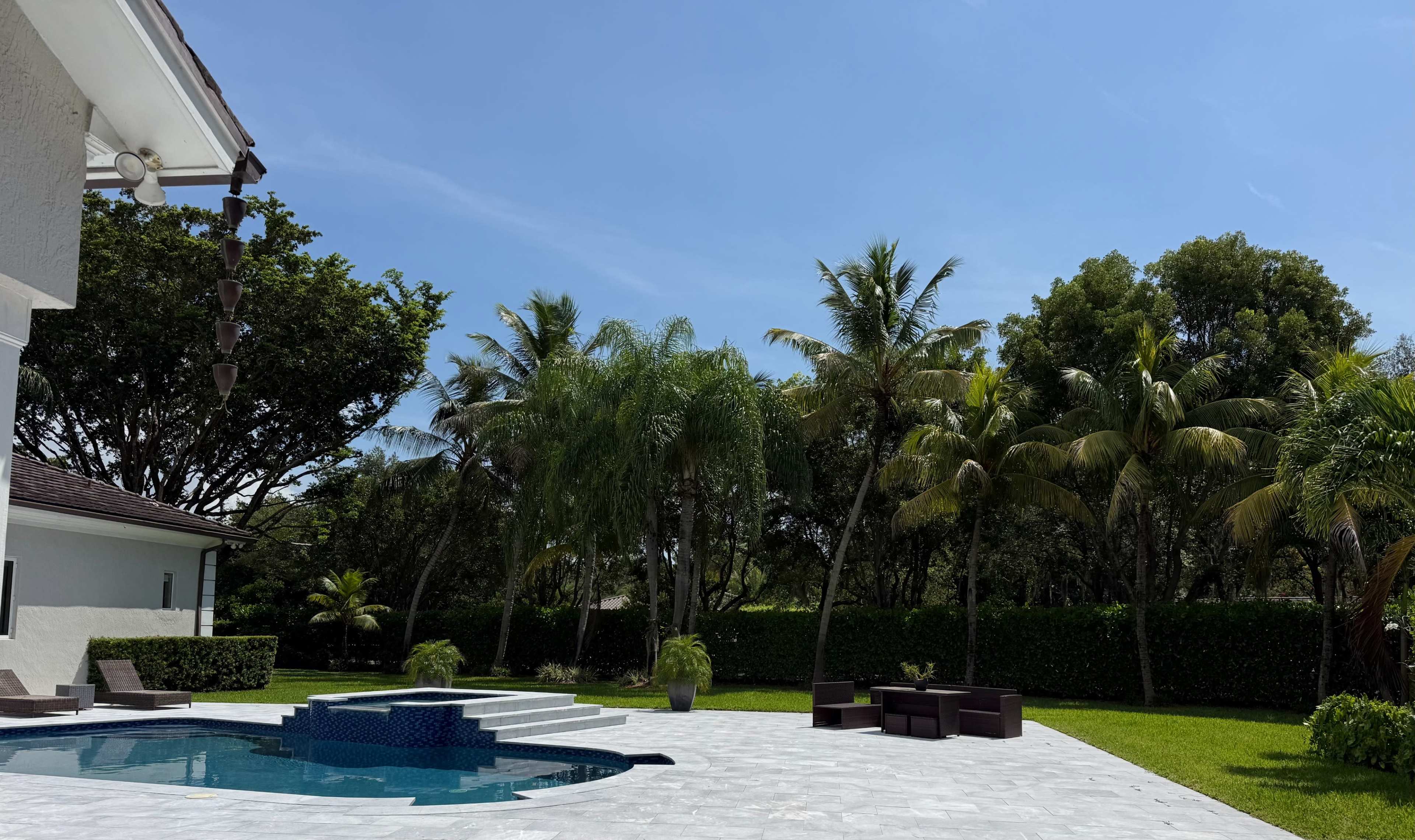 The image shows a backyard with a swimming pool surrounded by tropical trees and patio furniture on a sunny day.