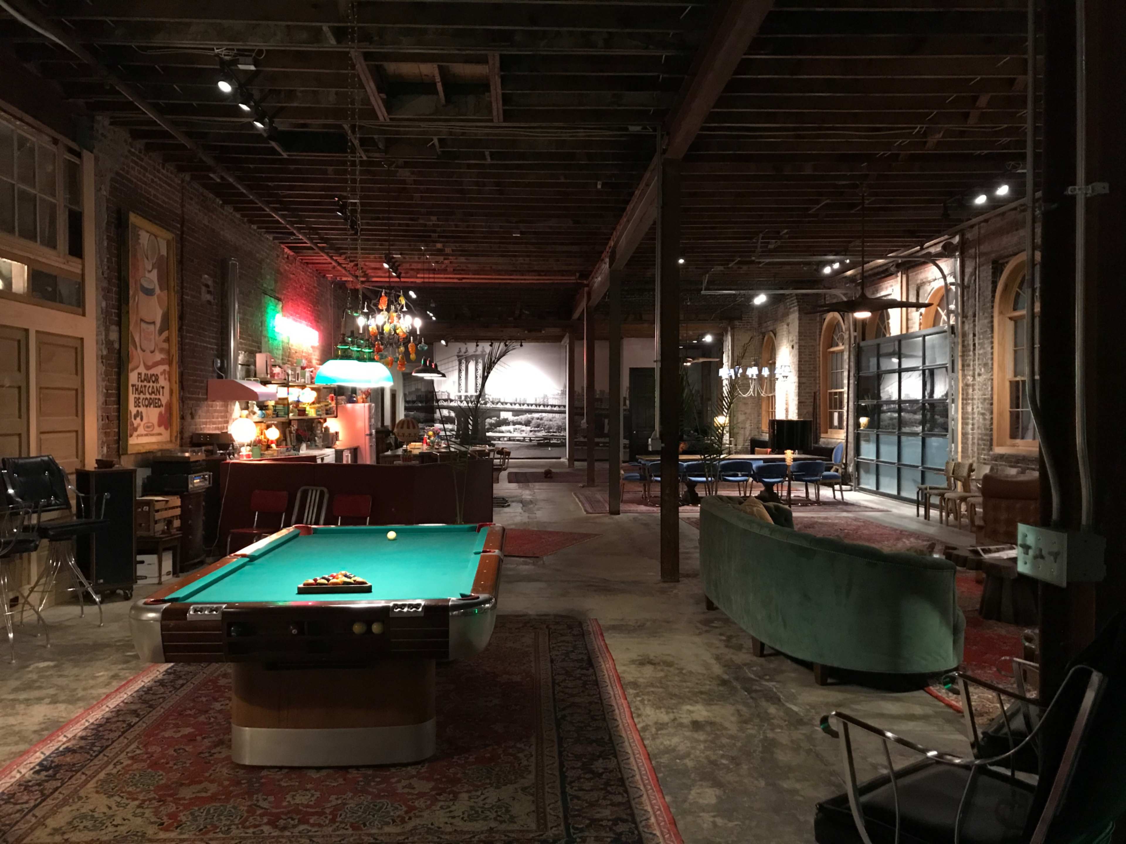 The image shows a spacious, industrial-style interior featuring a billiard table, vintage furniture, and exposed brick walls, illuminated by various light fixtures.