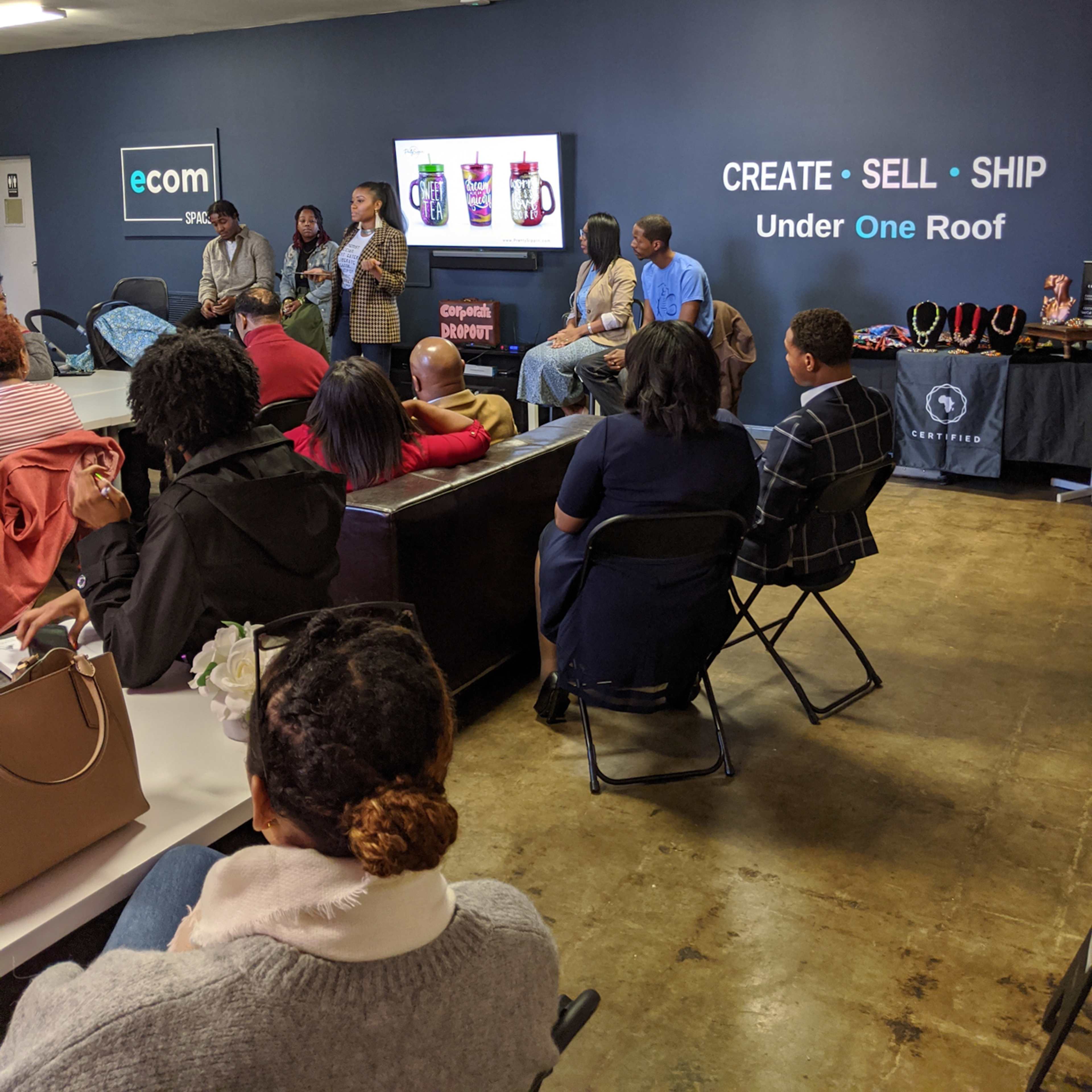 A group of people attends a presentation in a room with a screen displaying beverage containers and promotional materials.