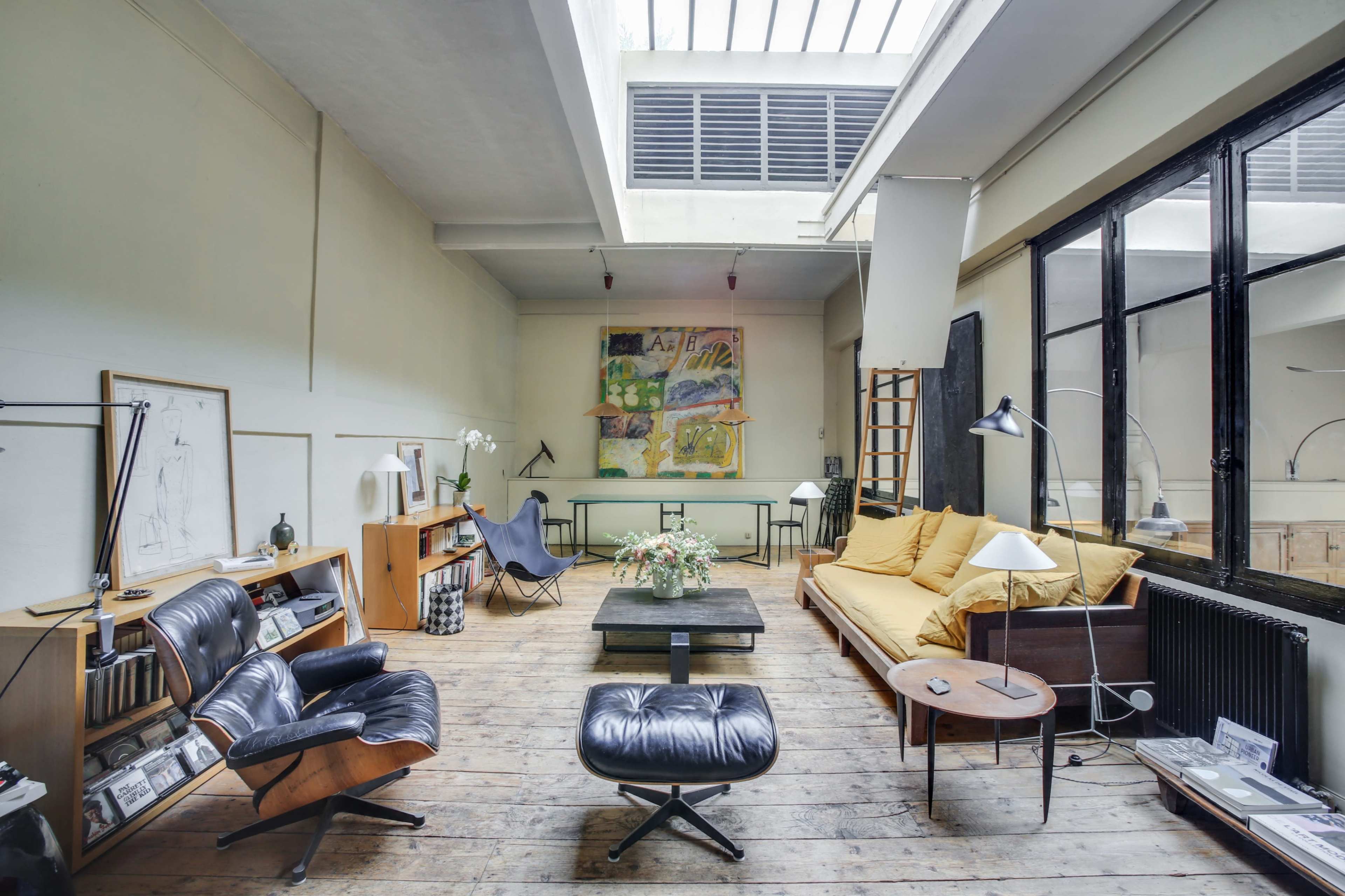 A spacious, modern living room with wooden flooring, large windows, a yellow sofa, and various pieces of furniture including an armchair and a coffee table.