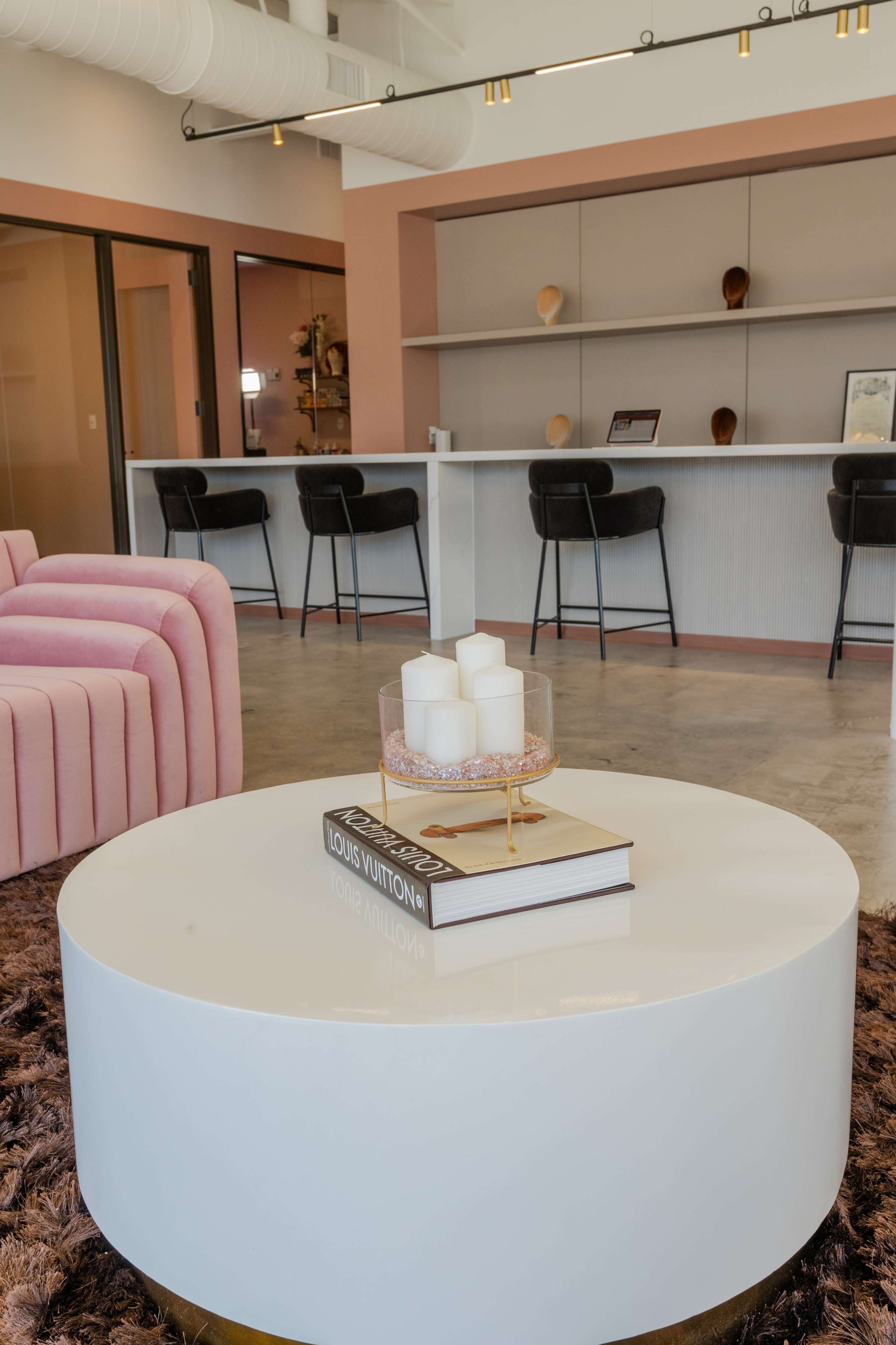 A modern lounge area features a circular coffee table with candles and books, surrounded by pink chairs and a bar counter in the background.