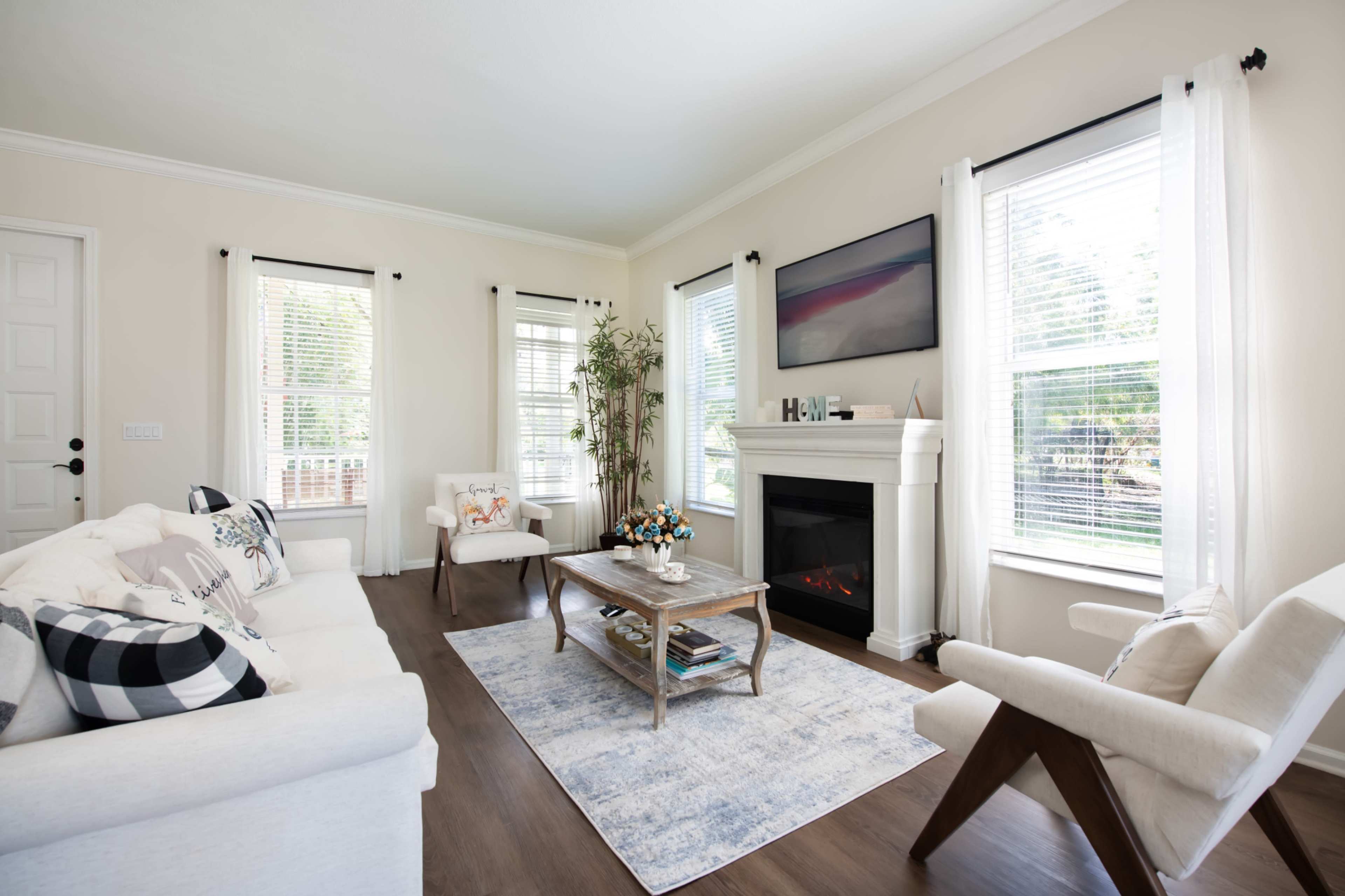A bright living room features a white sofa and armchair, a central coffee table, and a fireplace below a wall-mounted television, with large windows allowing natural light.