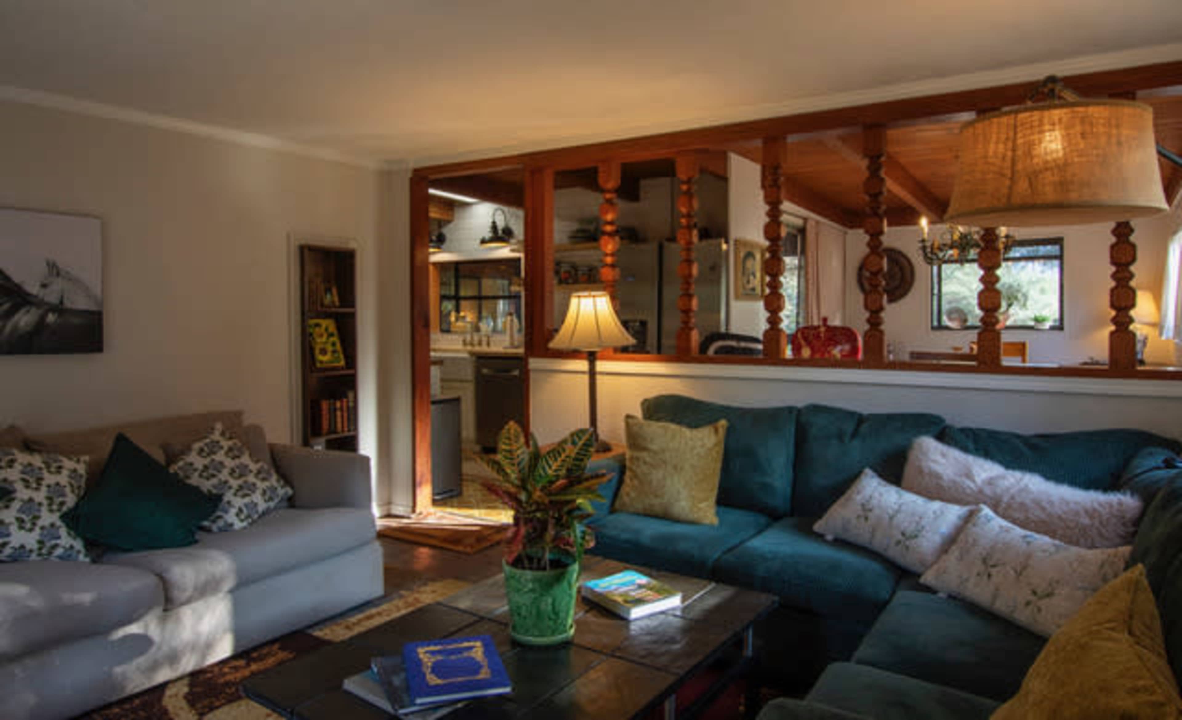 The image shows a cozy living room with teal and gray sofas, a wooden coffee table, and a view into a kitchen through a half-wall with decorative wooden posts.