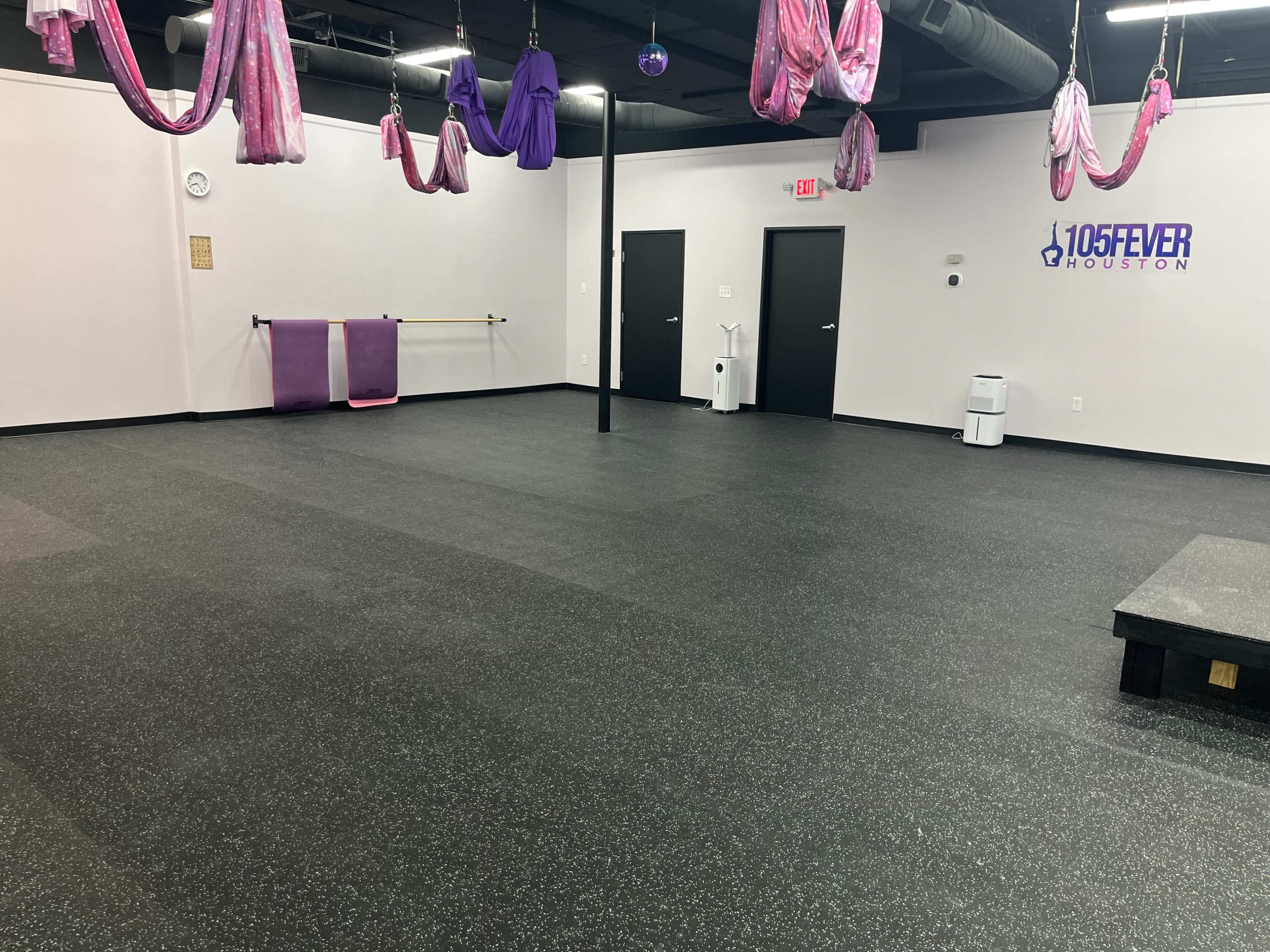 The image shows an empty fitness studio with black flooring, various purple fabrics hanging from the ceiling, and a stage area along one wall.