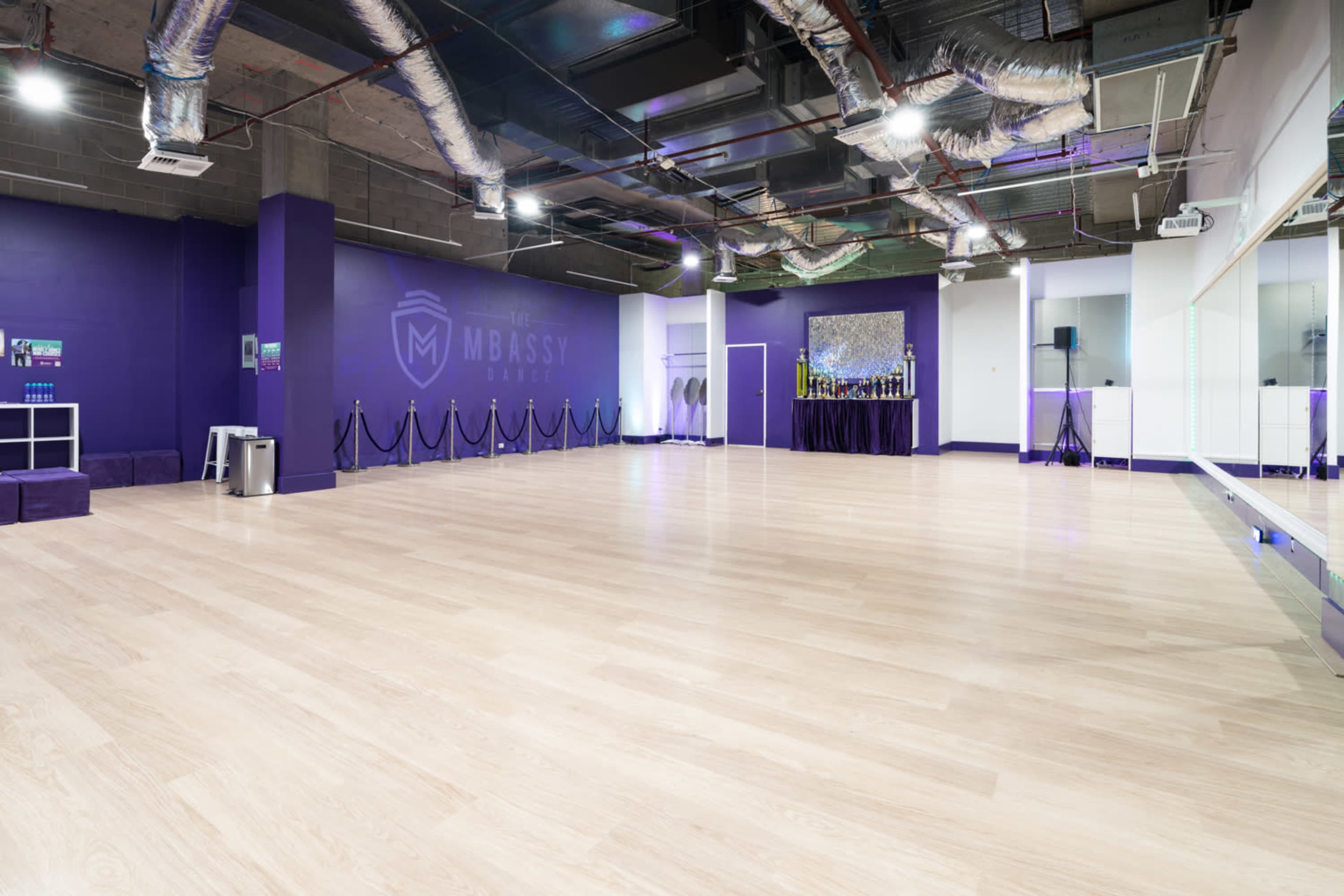 The image shows a spacious dance studio with a light wooden floor, purple walls, and mirrored panels.