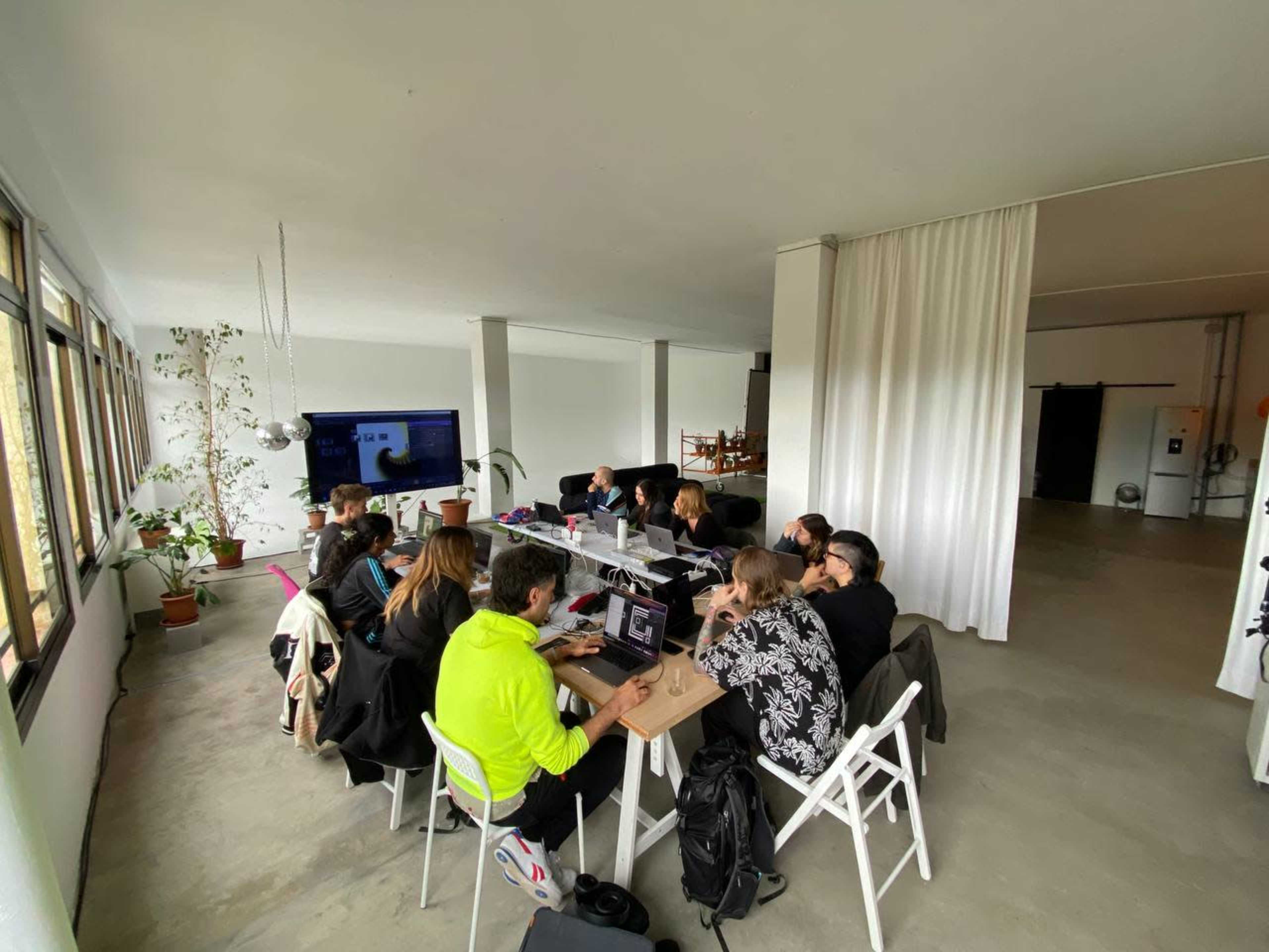 A group of people is seated around a long table in a bright, open room working on laptops.