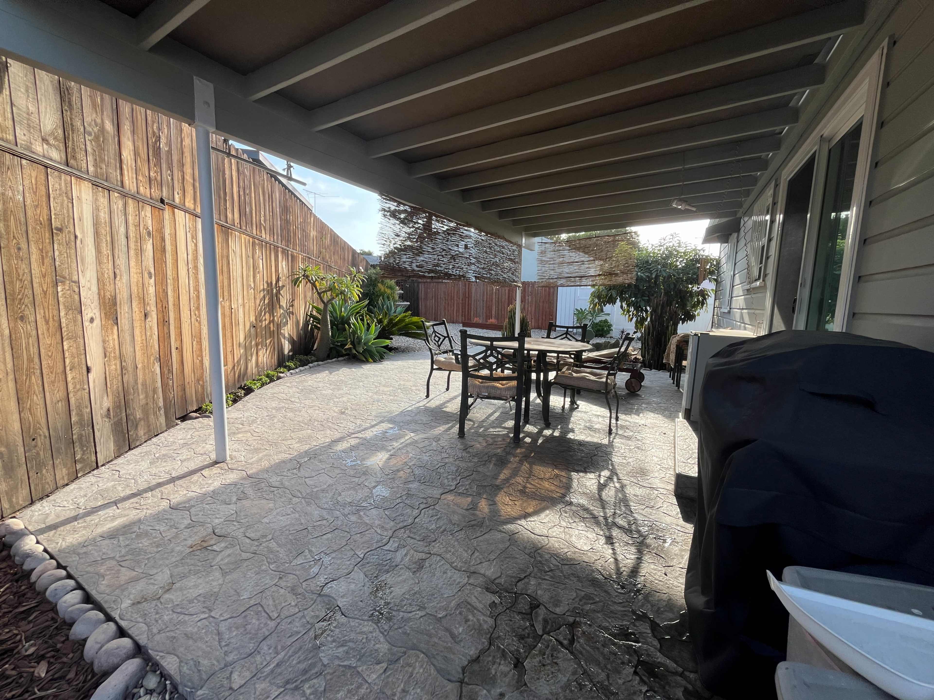 The image shows a covered patio area with a table and chairs, surrounded by wooden fencing and greenery.