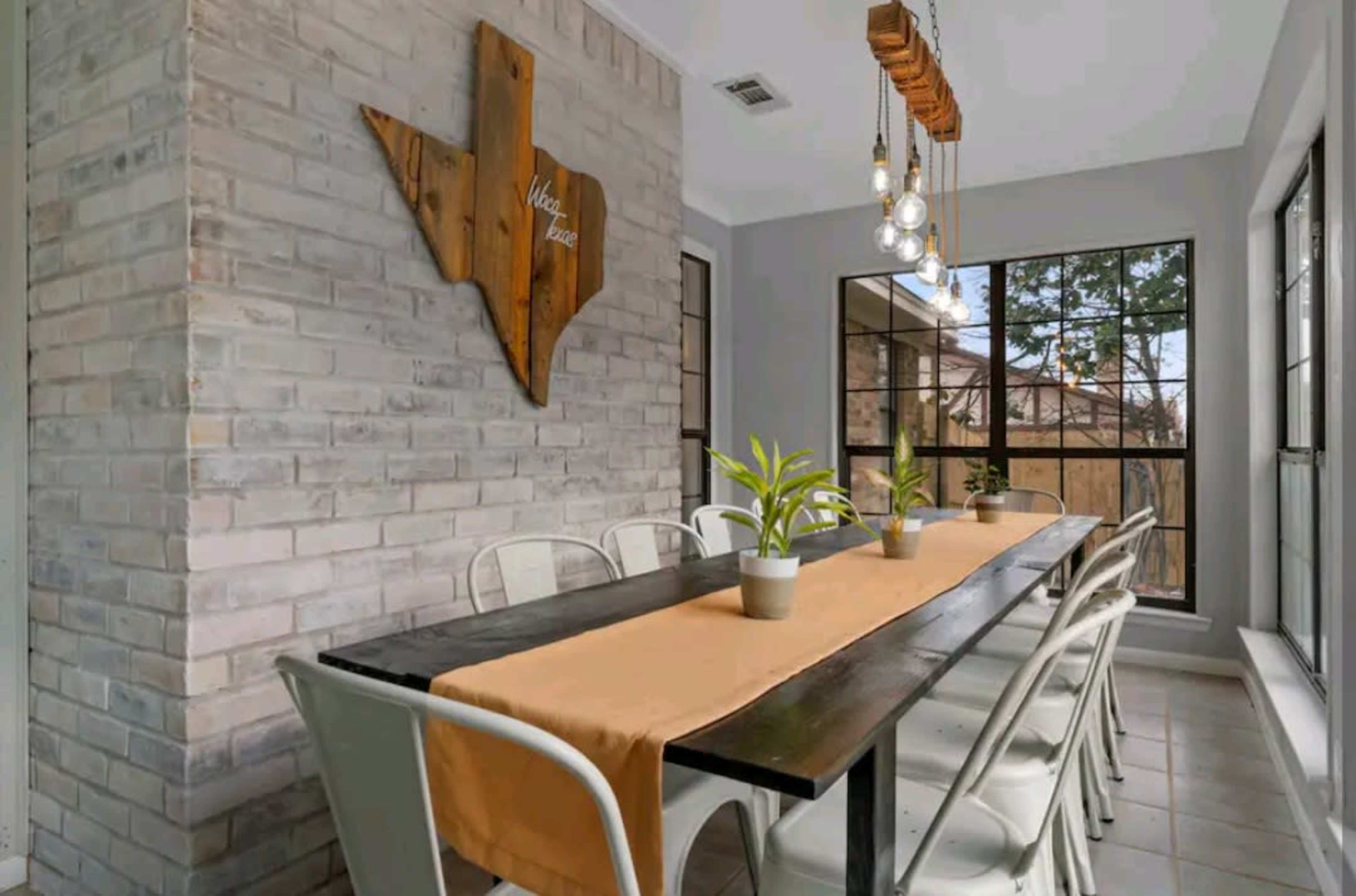 A dining area features a long table with white chairs, a beige table runner, and a wooden Texas-shaped wall decoration.