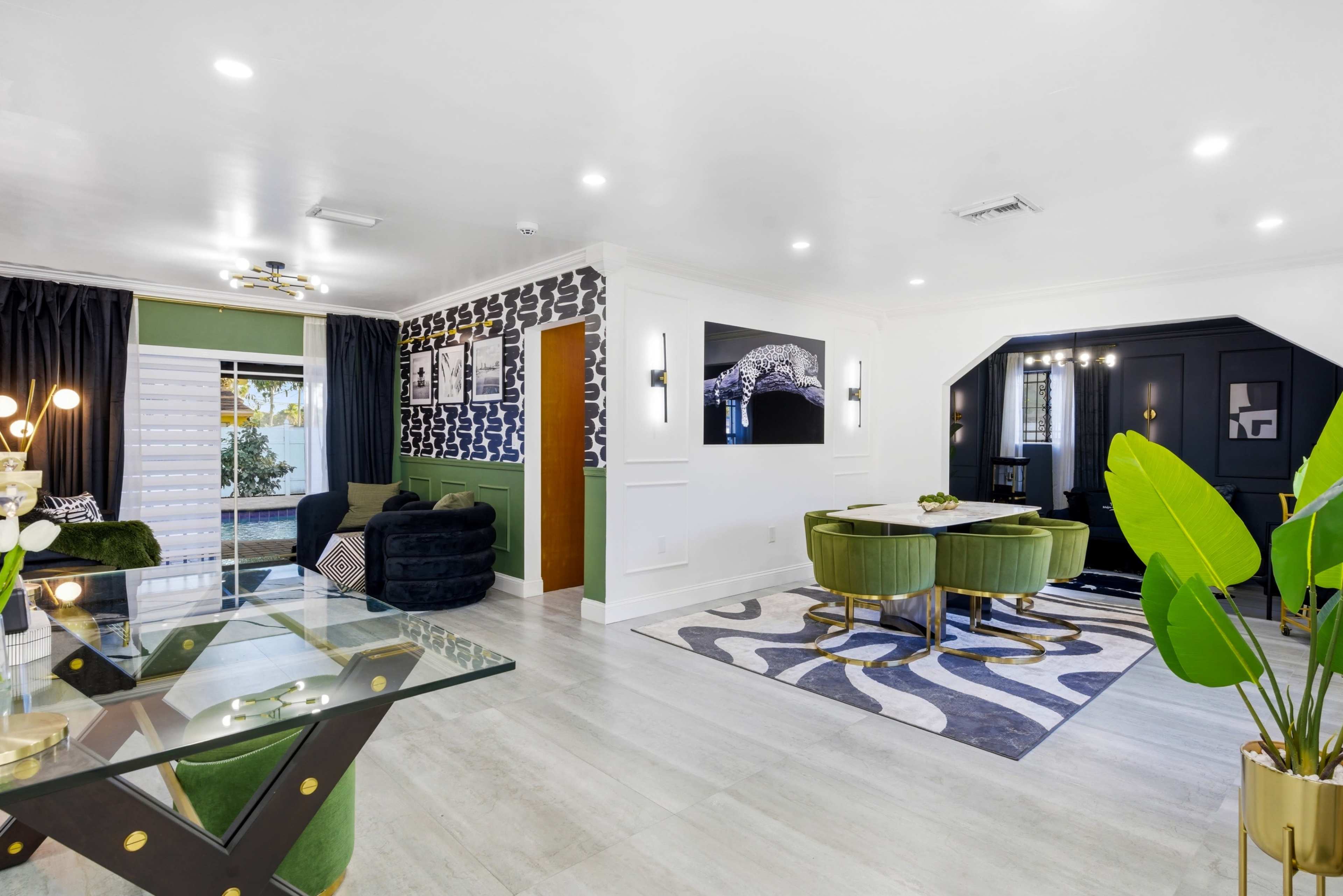 The image shows a modern living space with a glass table, green velvet chairs, dark walls featuring art, and a mix of decorative plants.