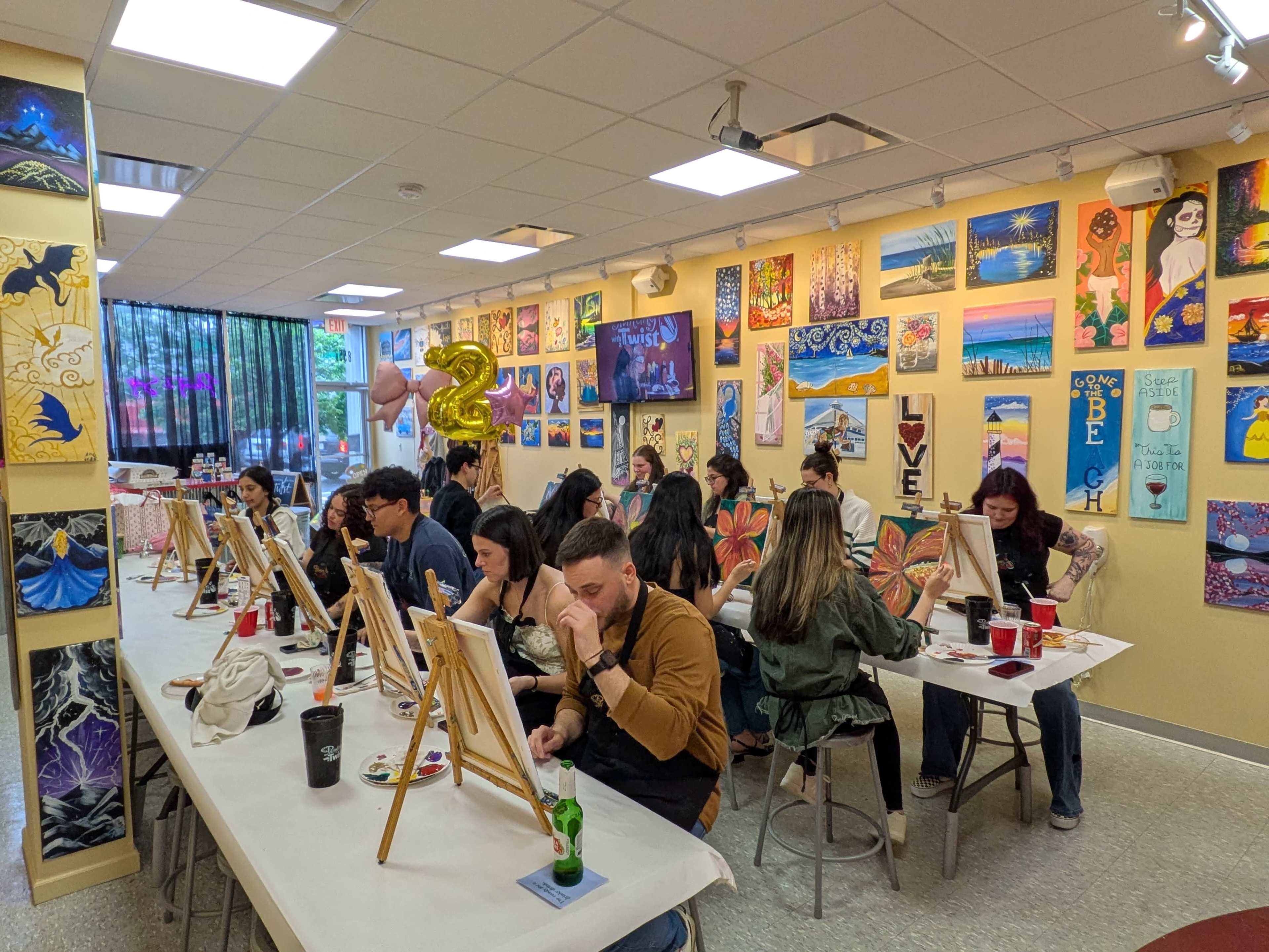 A group of people are painting on canvas at tables in a colorful art studio decorated with various artwork on the walls.