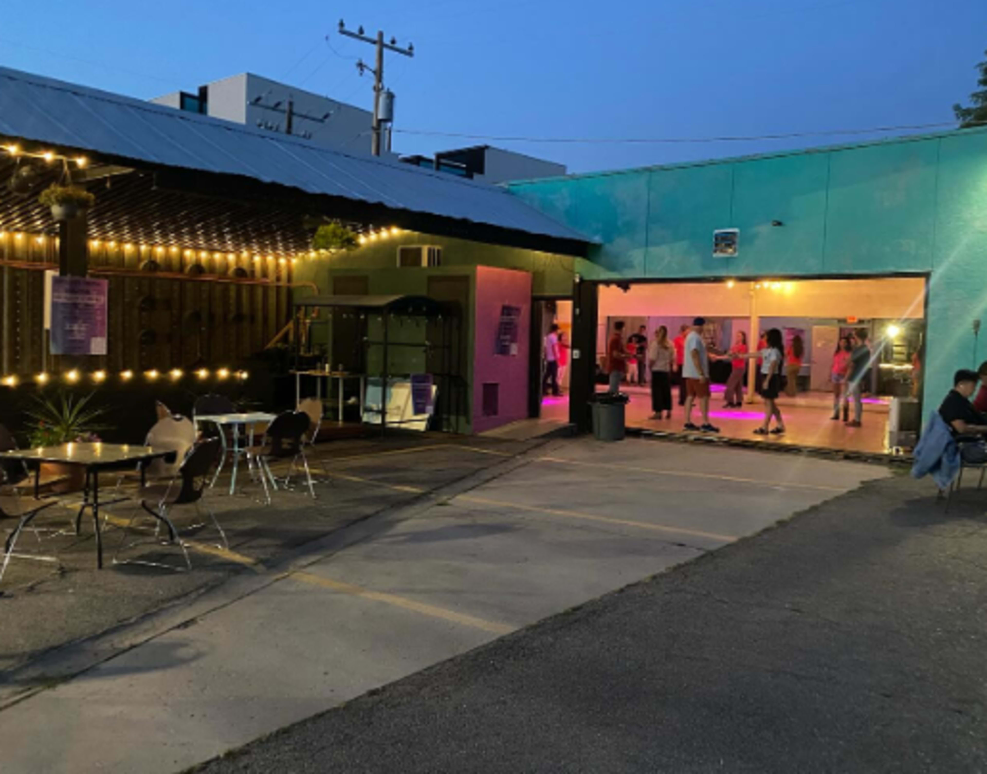The image shows a lively outdoor nightlife scene featuring people dancing in a vibrant venue with colorful lighting, while tables and chairs are arranged nearby.