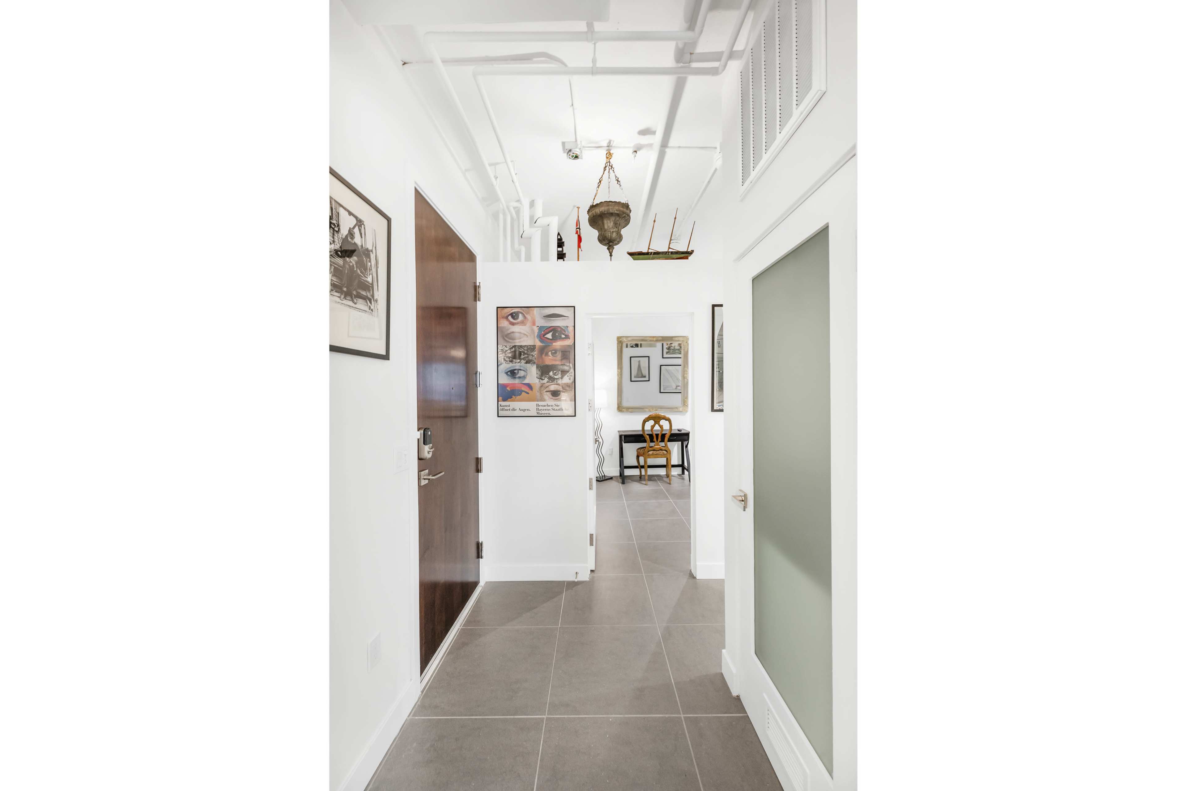 A hallway with white walls and tiled flooring leads to a small seating area, featuring artwork and a decorative chandelier.