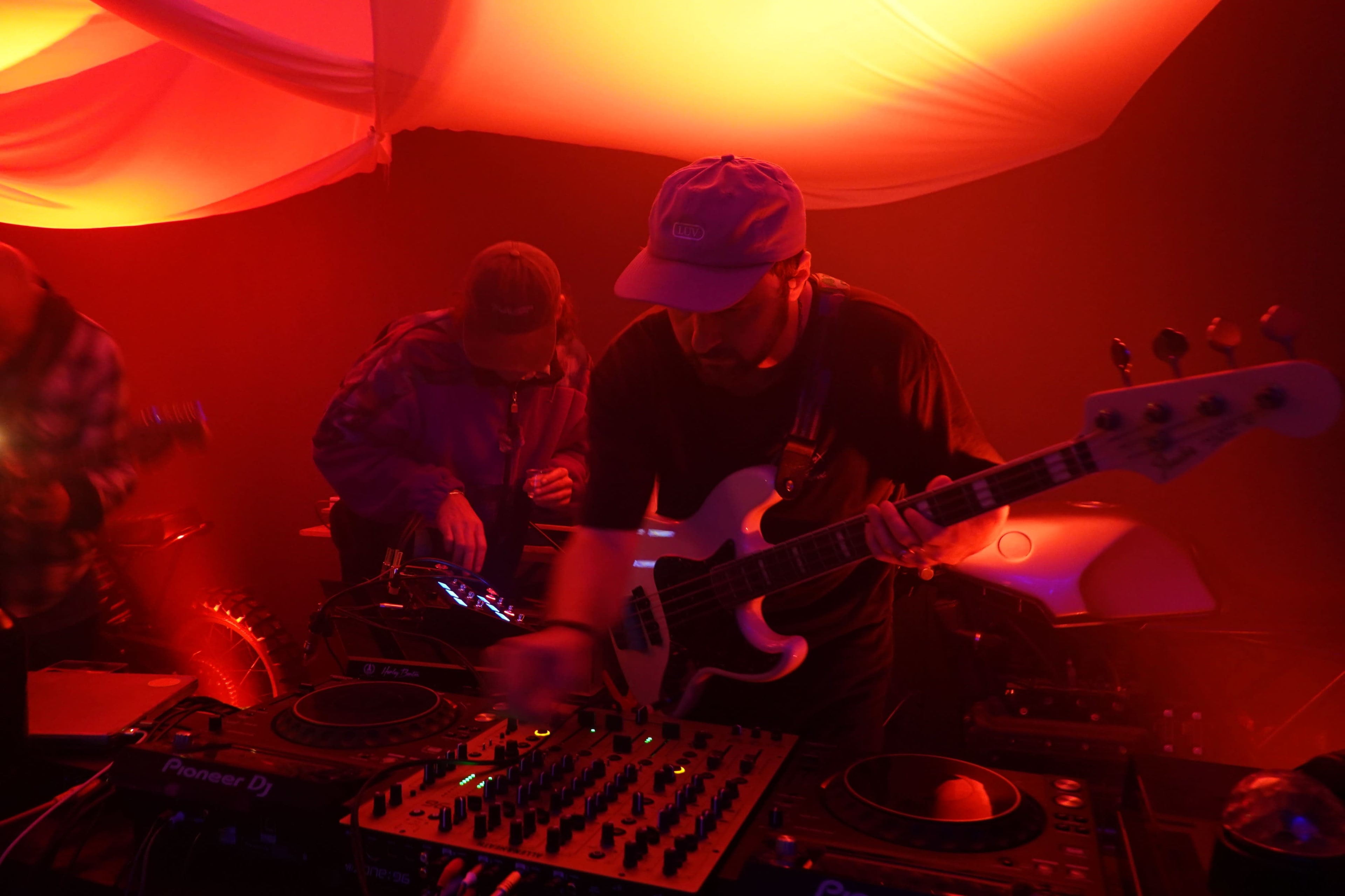 A musician plays bass guitar and adjusts equipment at a dimly lit performance space with colored lighting.