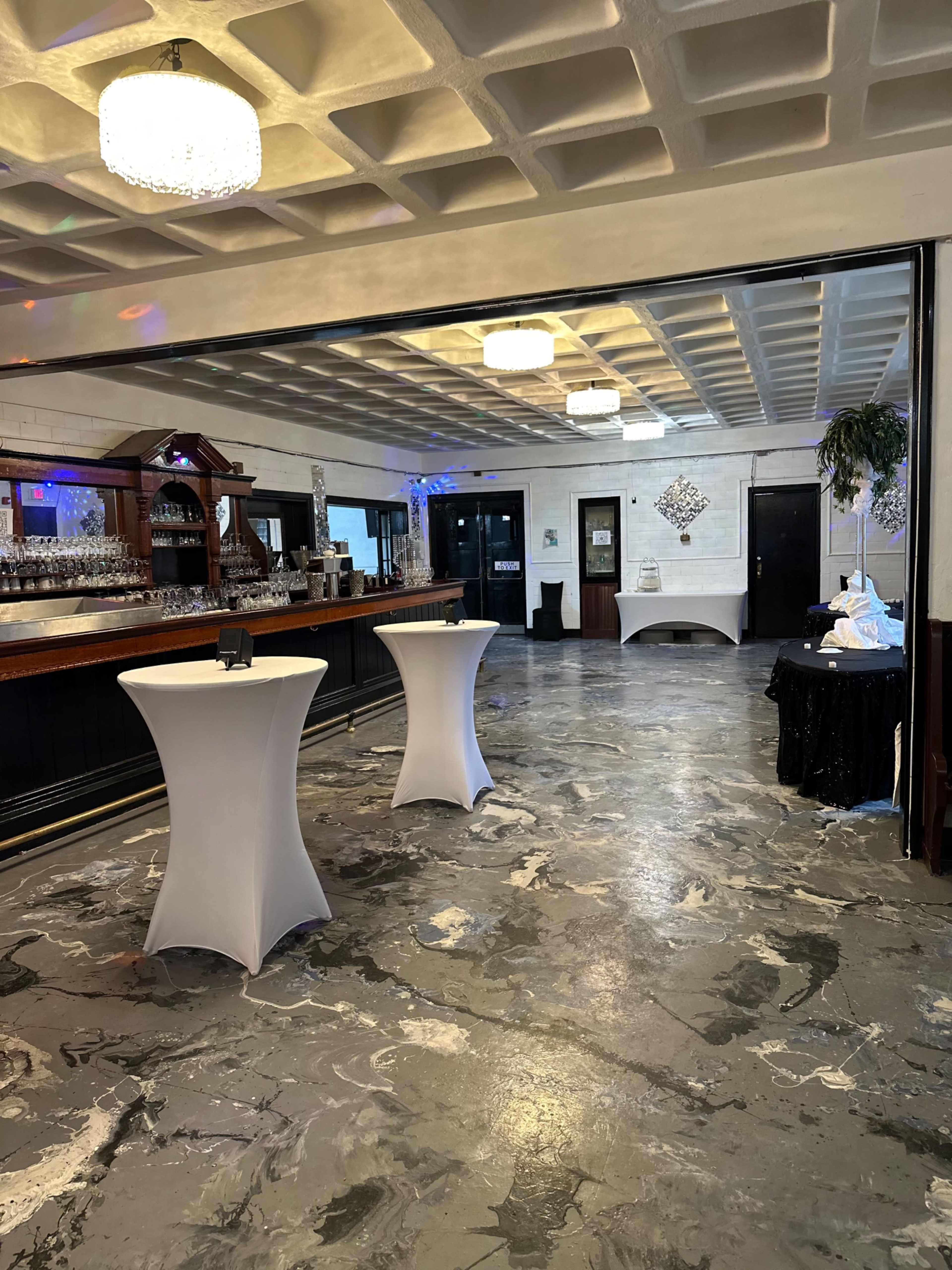 The interior of a venue features a polished concrete floor, a long bar with glassware, and several high tables set up in the space.