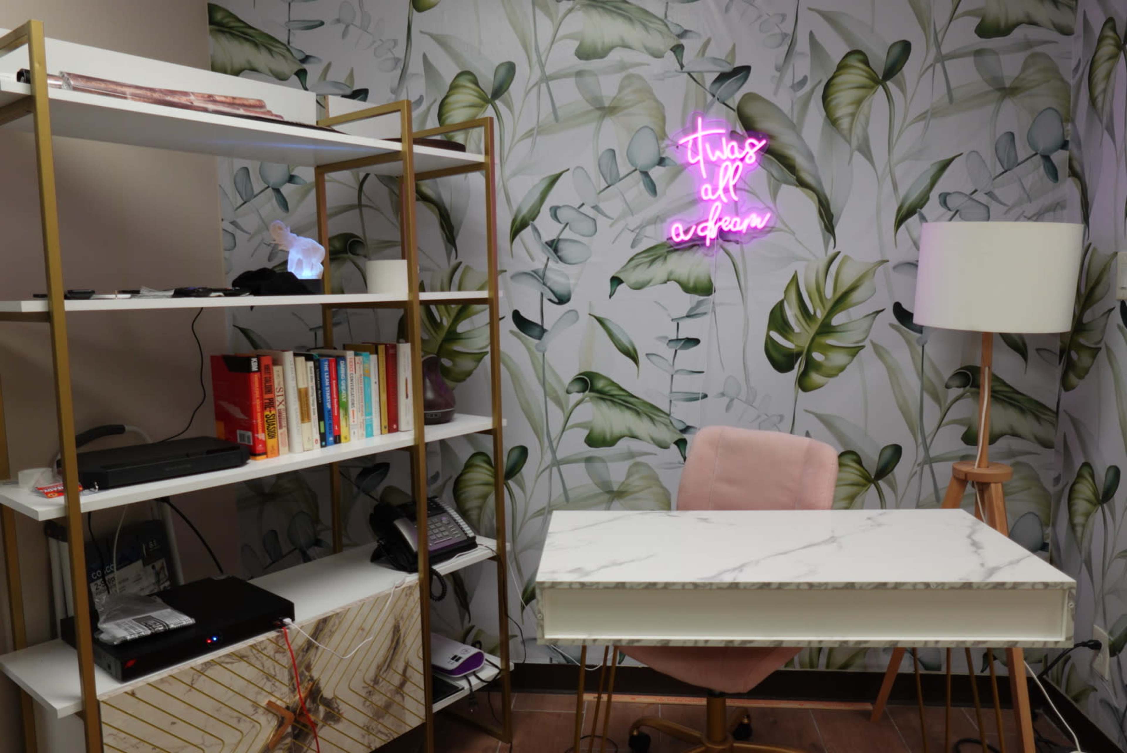 A modern office space features a marble-top desk, a bookshelf filled with books, and a neon sign on a leaf-patterned wall.